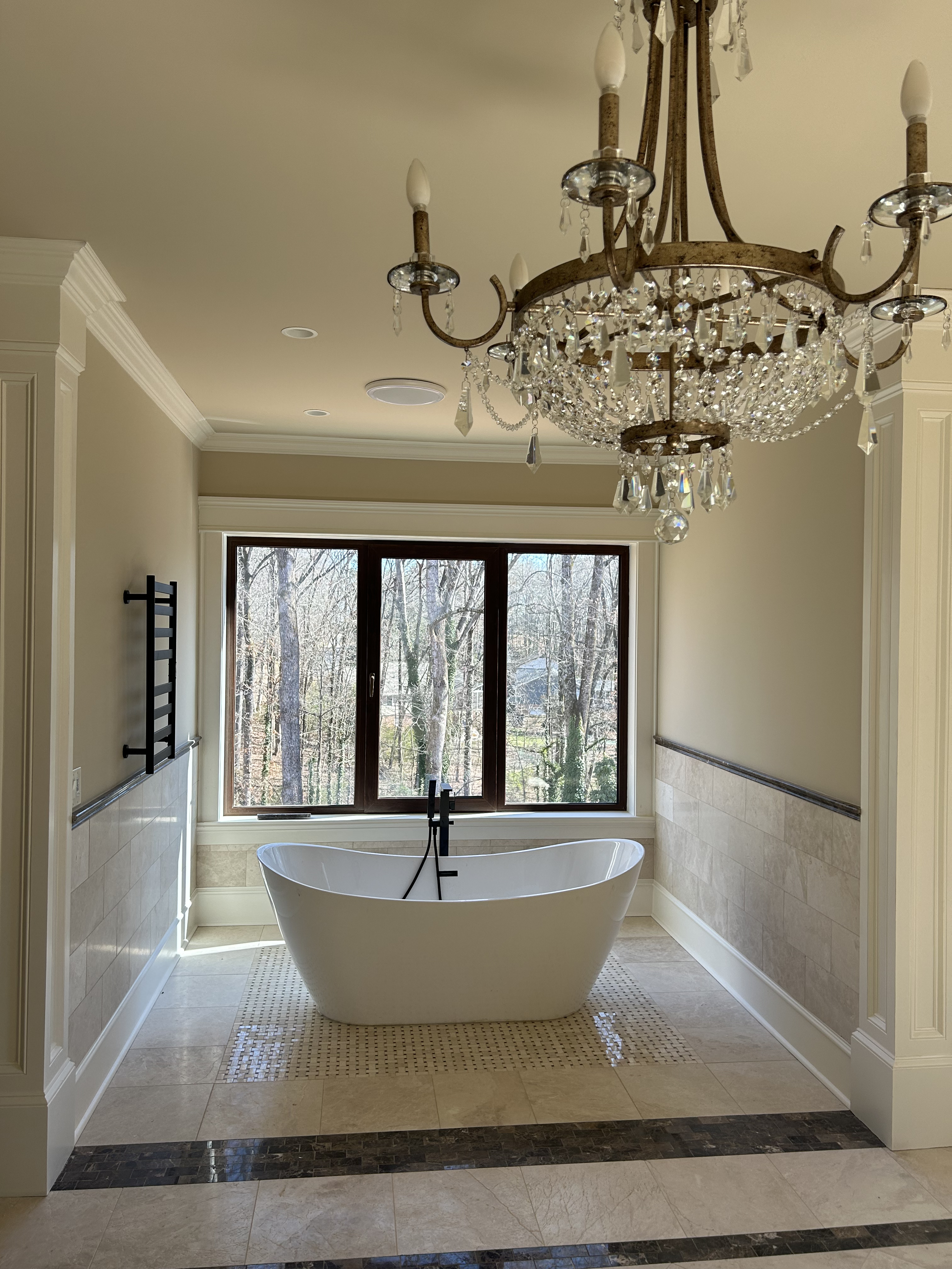 Traditional luxury bathroom with freestanding soaking tub at window, marble wainscoting, brass fixtures, crystal chandelier, heated rack