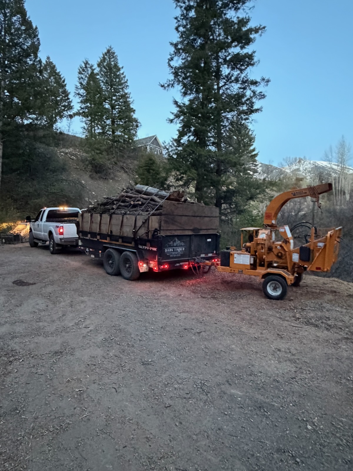 Truck with loaded Dark Timber trailer and orange wood chipper at dusk with mountain backdrop