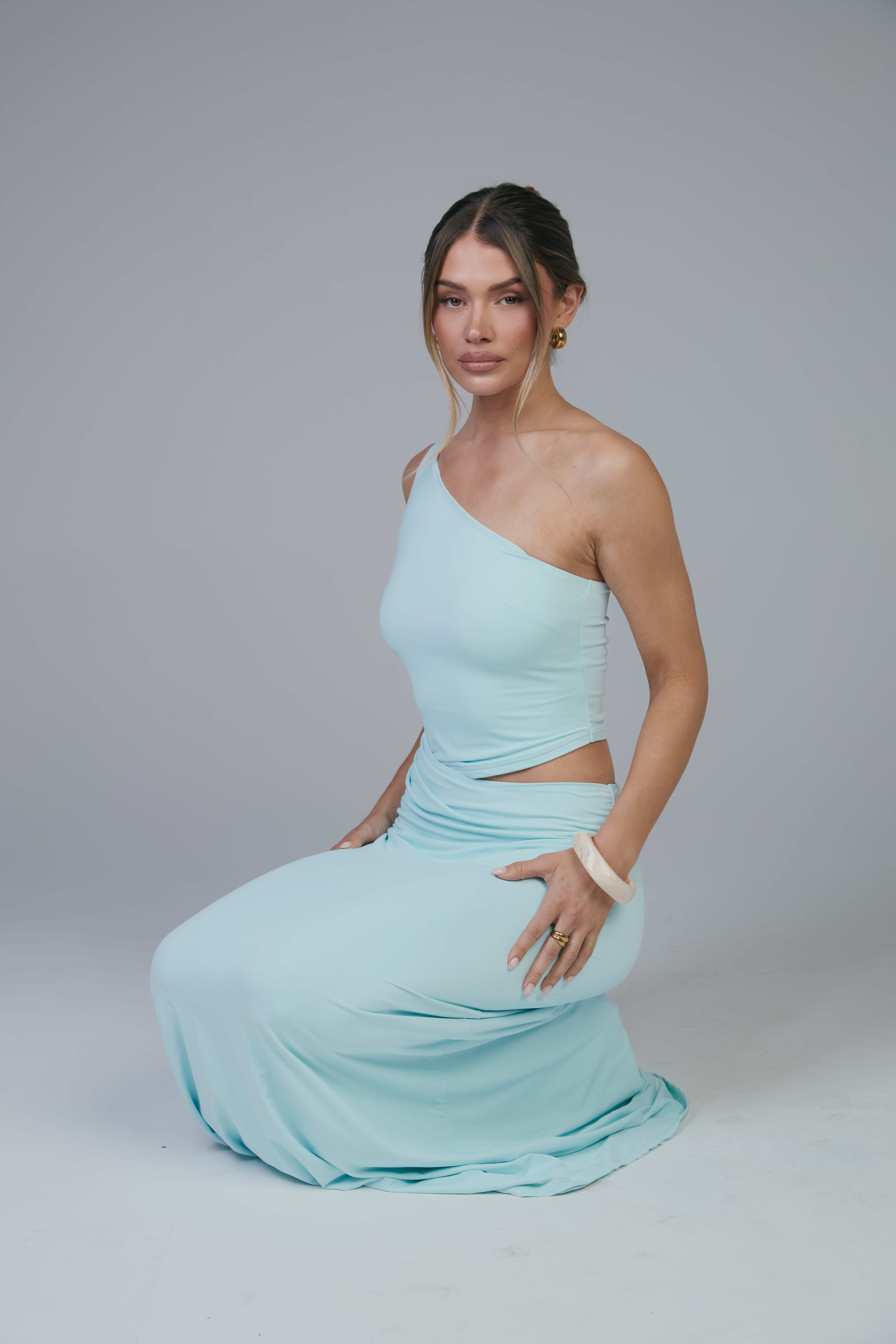Mollie kneeling in aqua one-shoulder top and maxi skirt with hand on hip