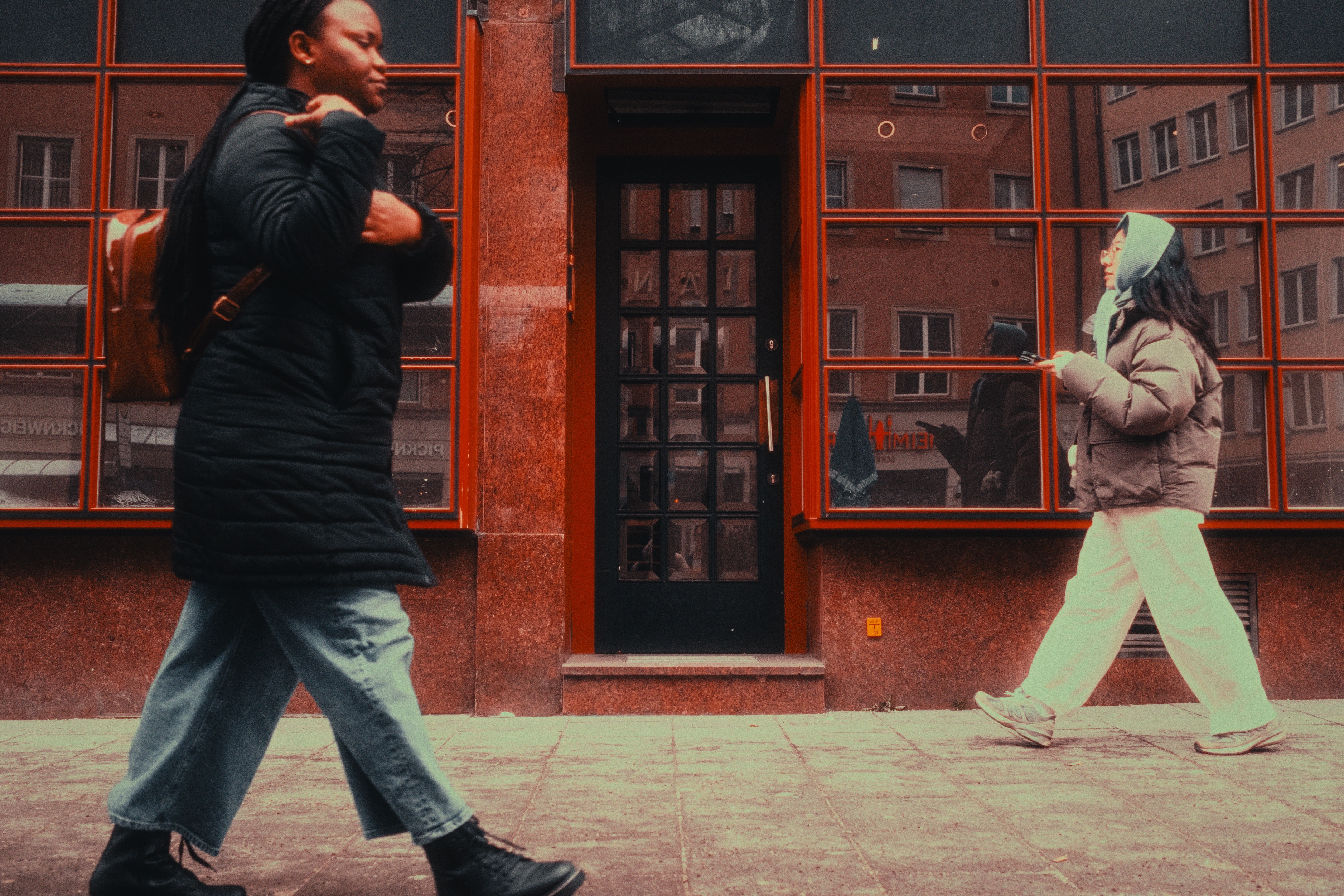 Two pedestrians crossing past a red-framed glass storefront in Munich