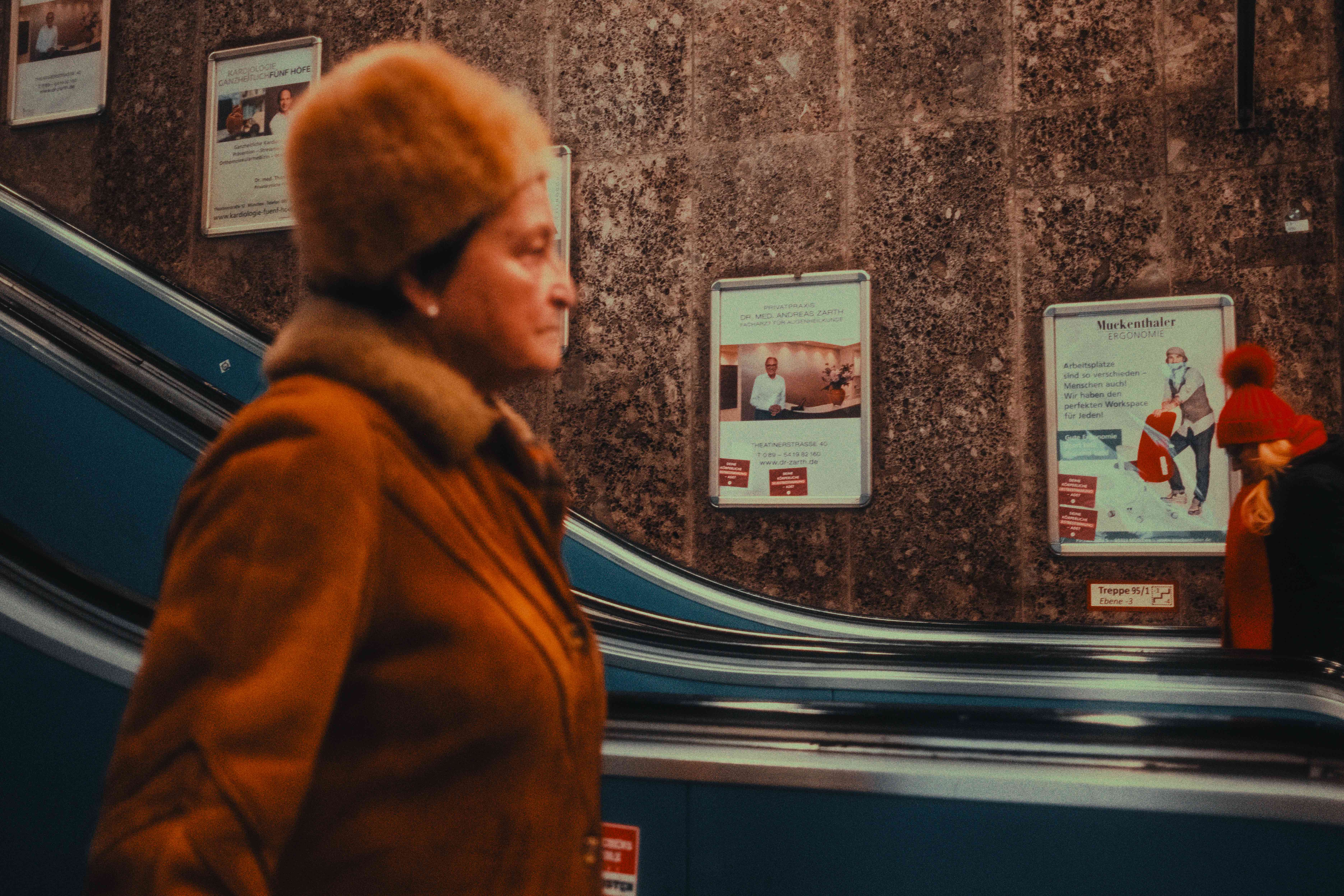 Older woman in an ochre fur-collar coat riding an escalator past transit posters