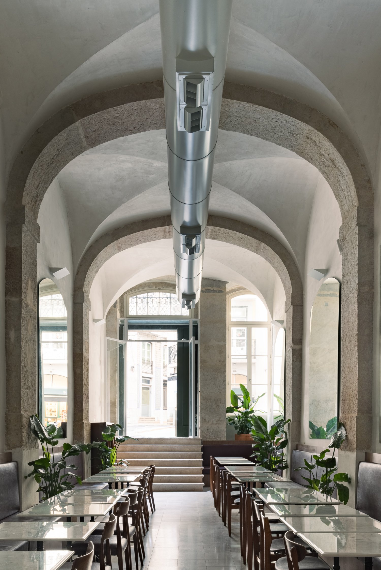 Elegant dining corridor with vaulted ceilings, stone arches, and rows of glass-top tables, featuring abundant natural light and tropical plants