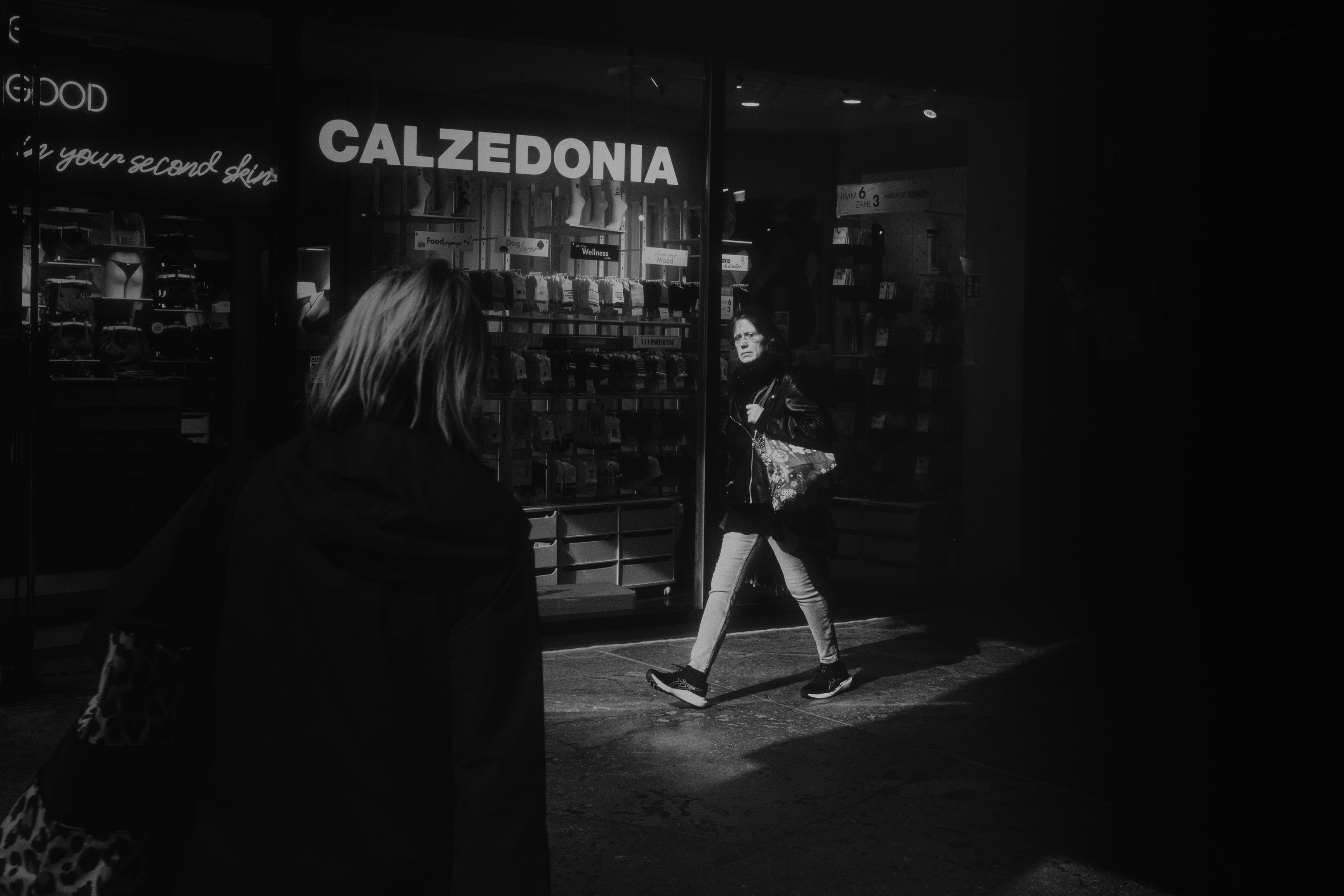 High-contrast black and white night frame of a woman passing a lit storefront