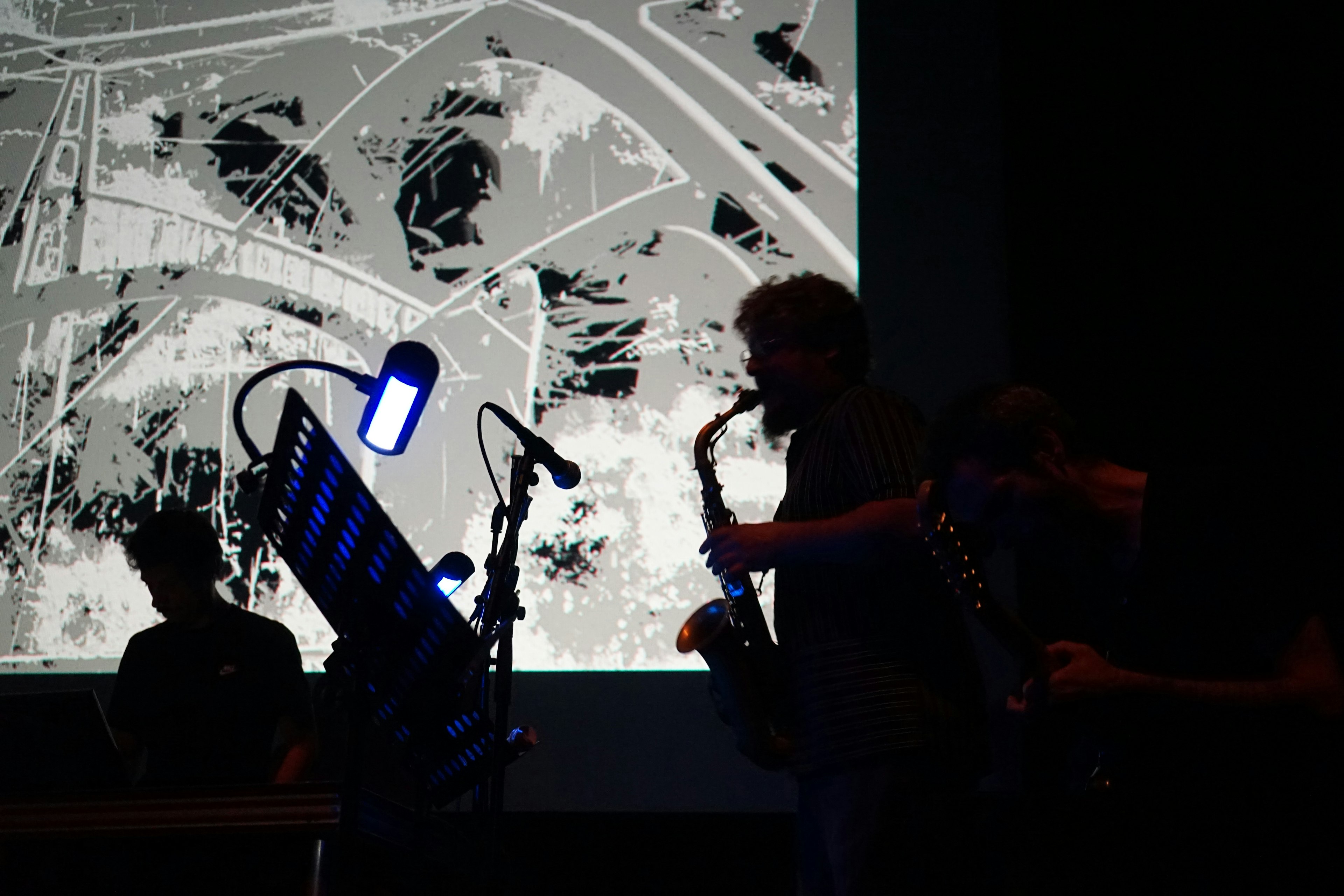 Live performance of On/Off showing silhouetted musicians with saxophone against projected abstract visuals