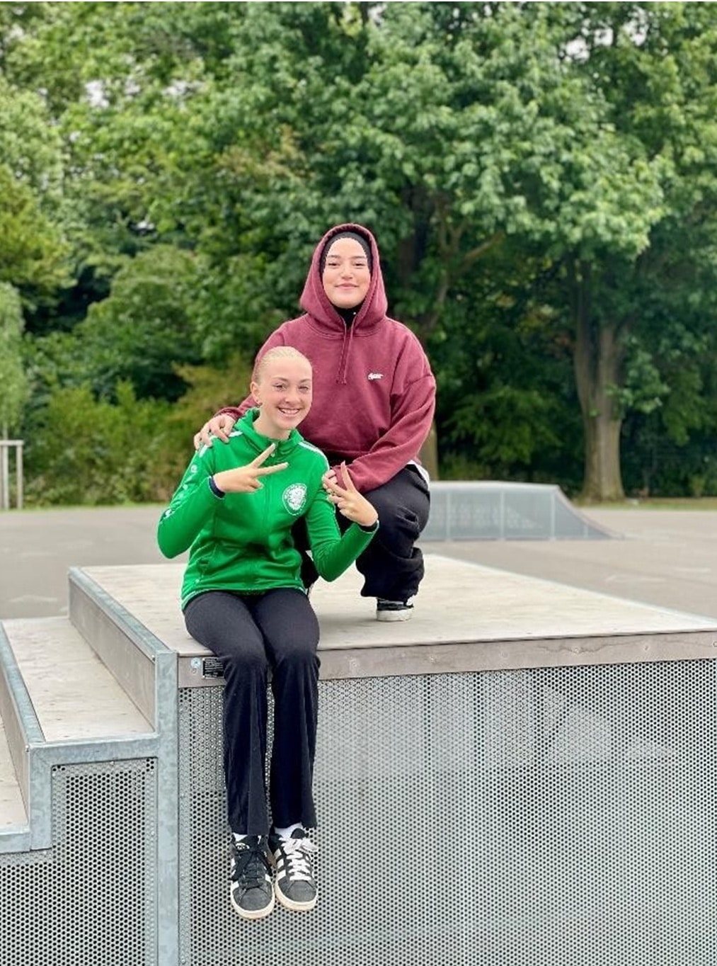 Zwei junge Frauen sitzen auf einem Skatepark-Element und lächeln in die Kamera