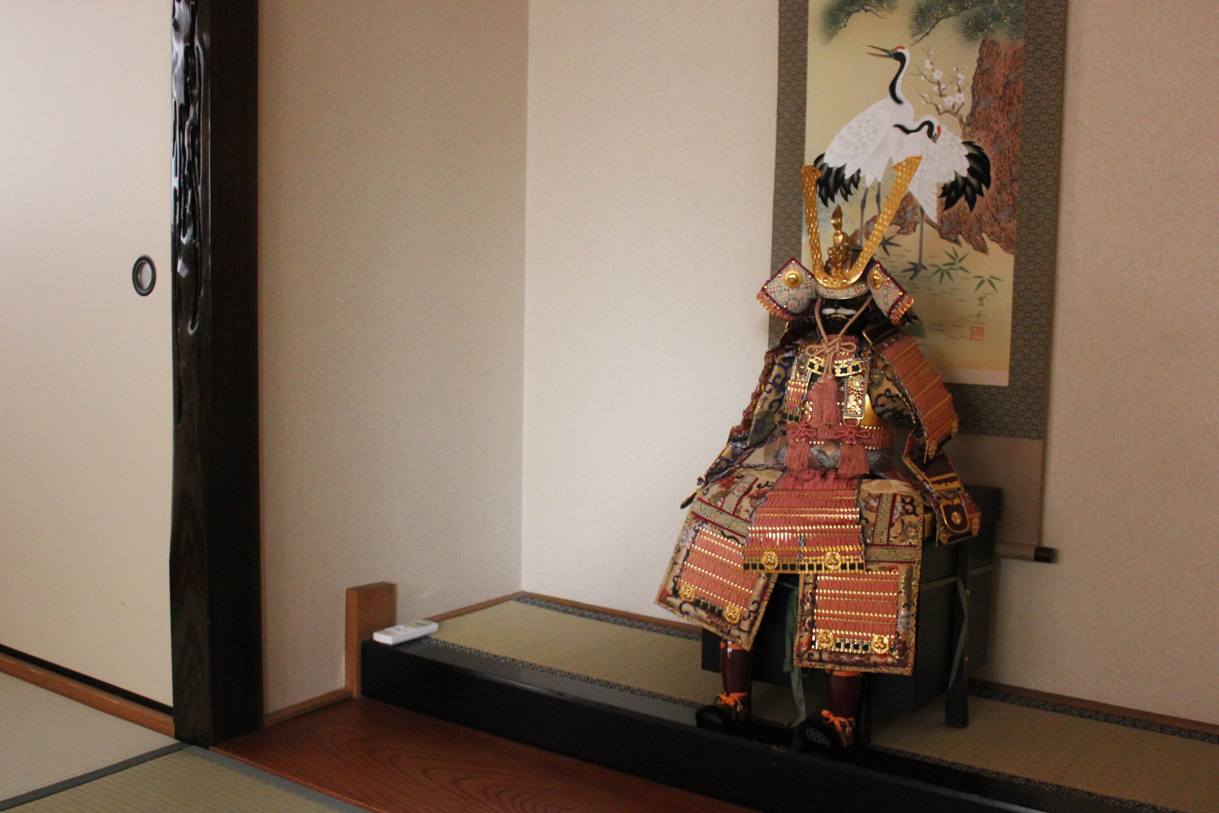 Traditional tokonoma alcove with ornate miniature samurai armor in red and gold brocade, gold-horned kabuto helmet, displayed on black lacquer platform with crane kakejiku scroll painting