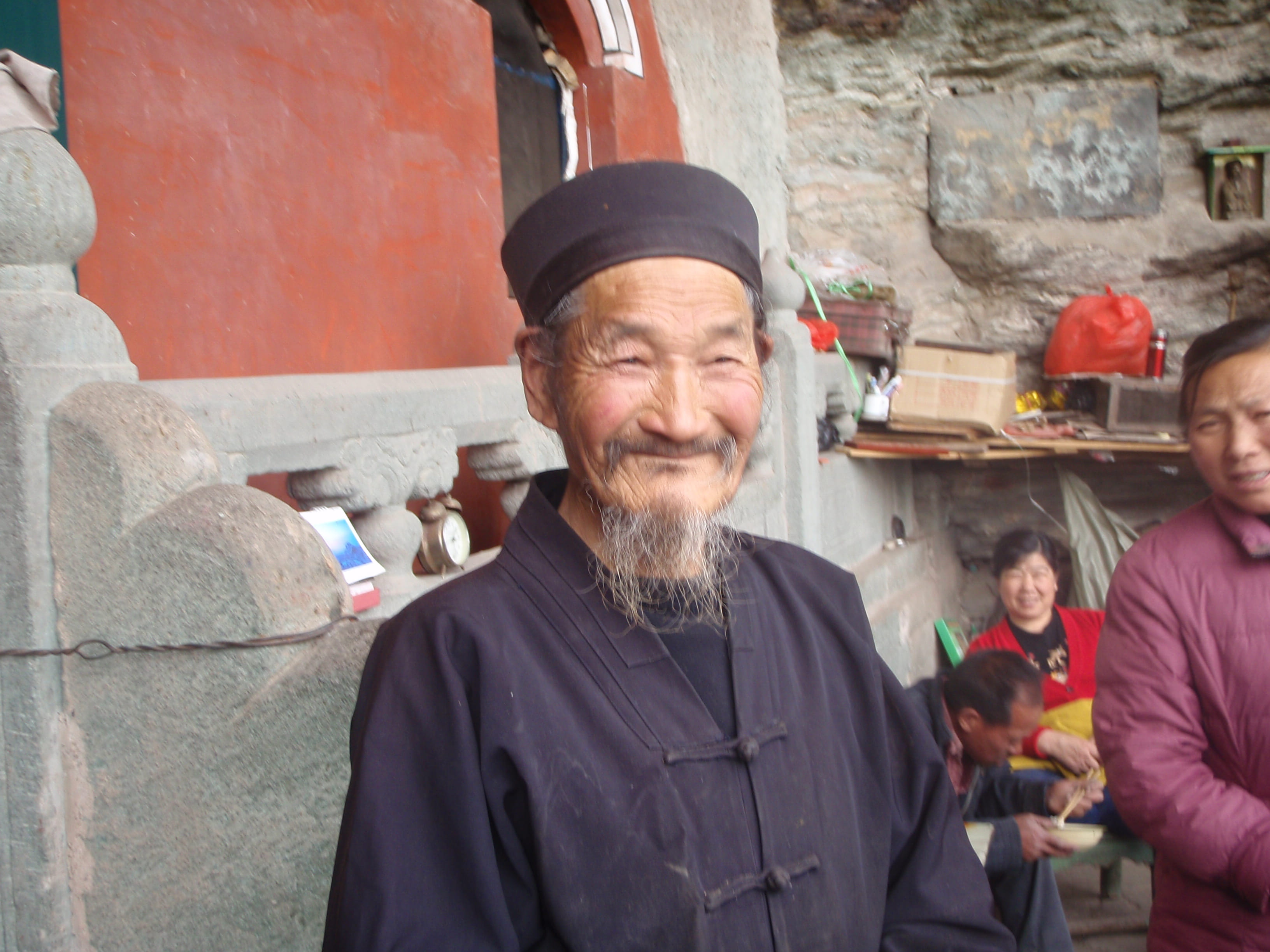 Elderly Chinese master with long white beard and traditional dark robe and cap, smiling joyfully outdoors with warm colorful background