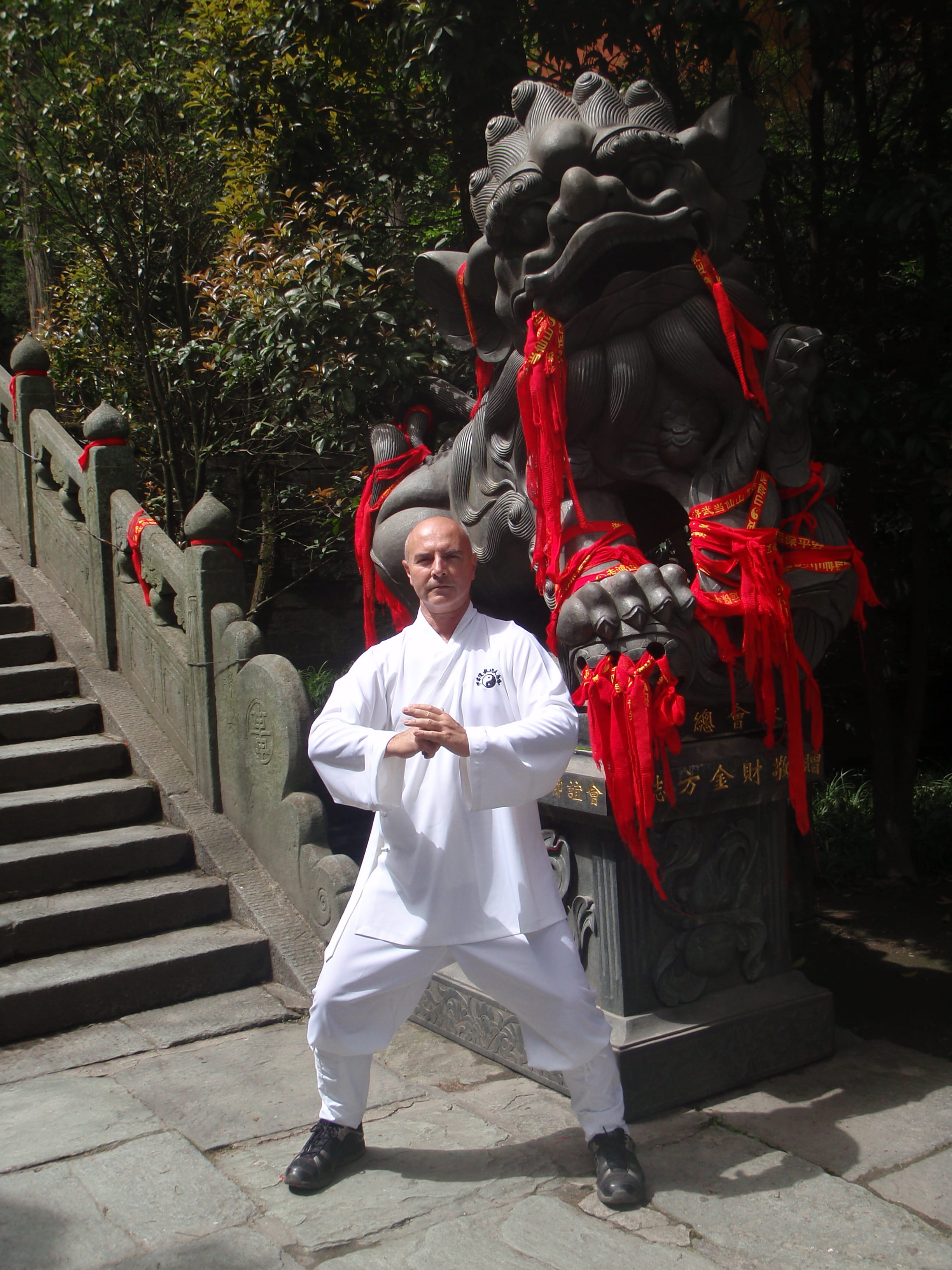 Kadama in a formal qigong stance in front of a large red-ribboned stone guardian statue at a Chinese temple