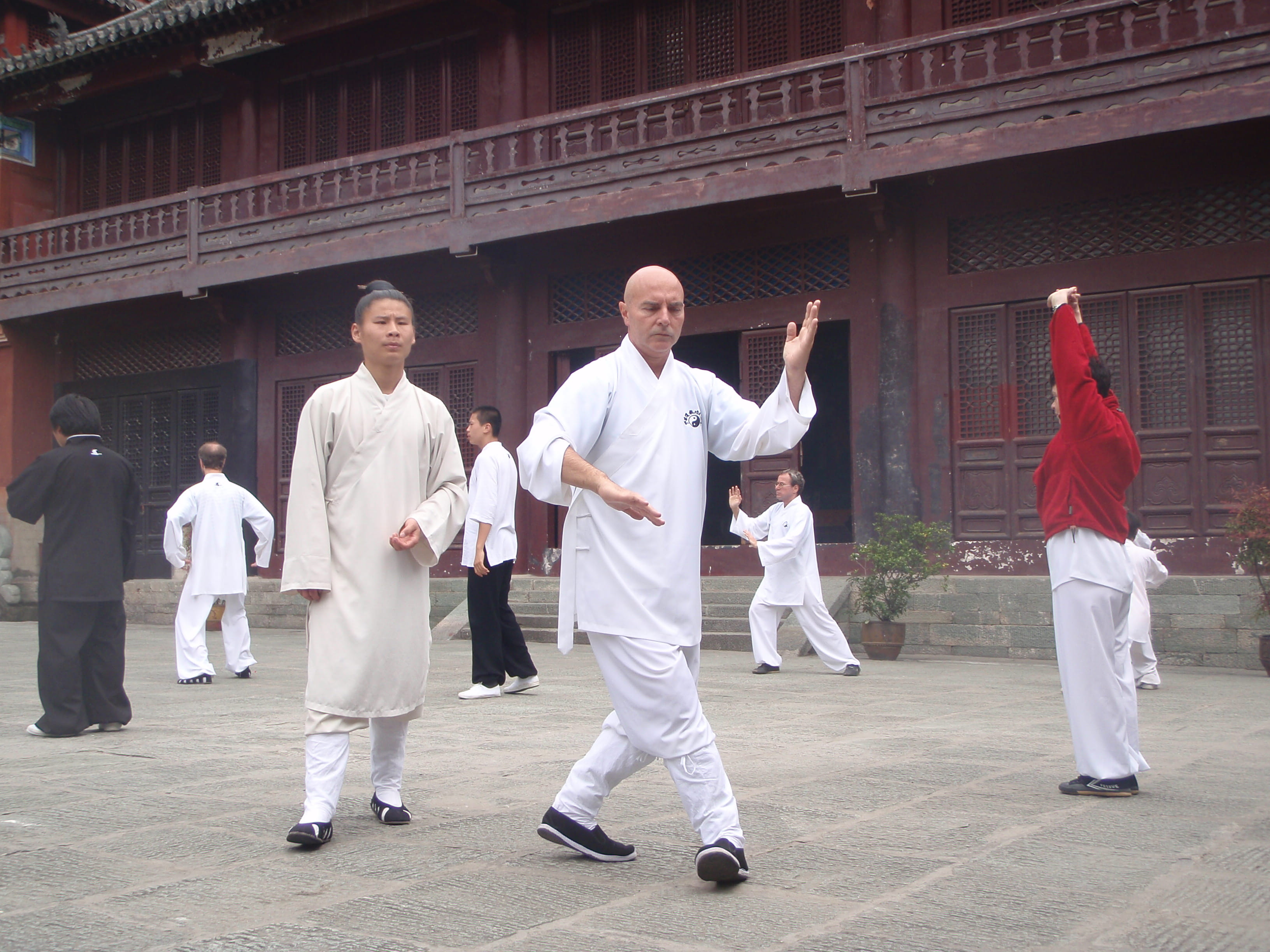 Kadama demonstrating a flowing forward qigong posture in a temple courtyard with female master observing at left