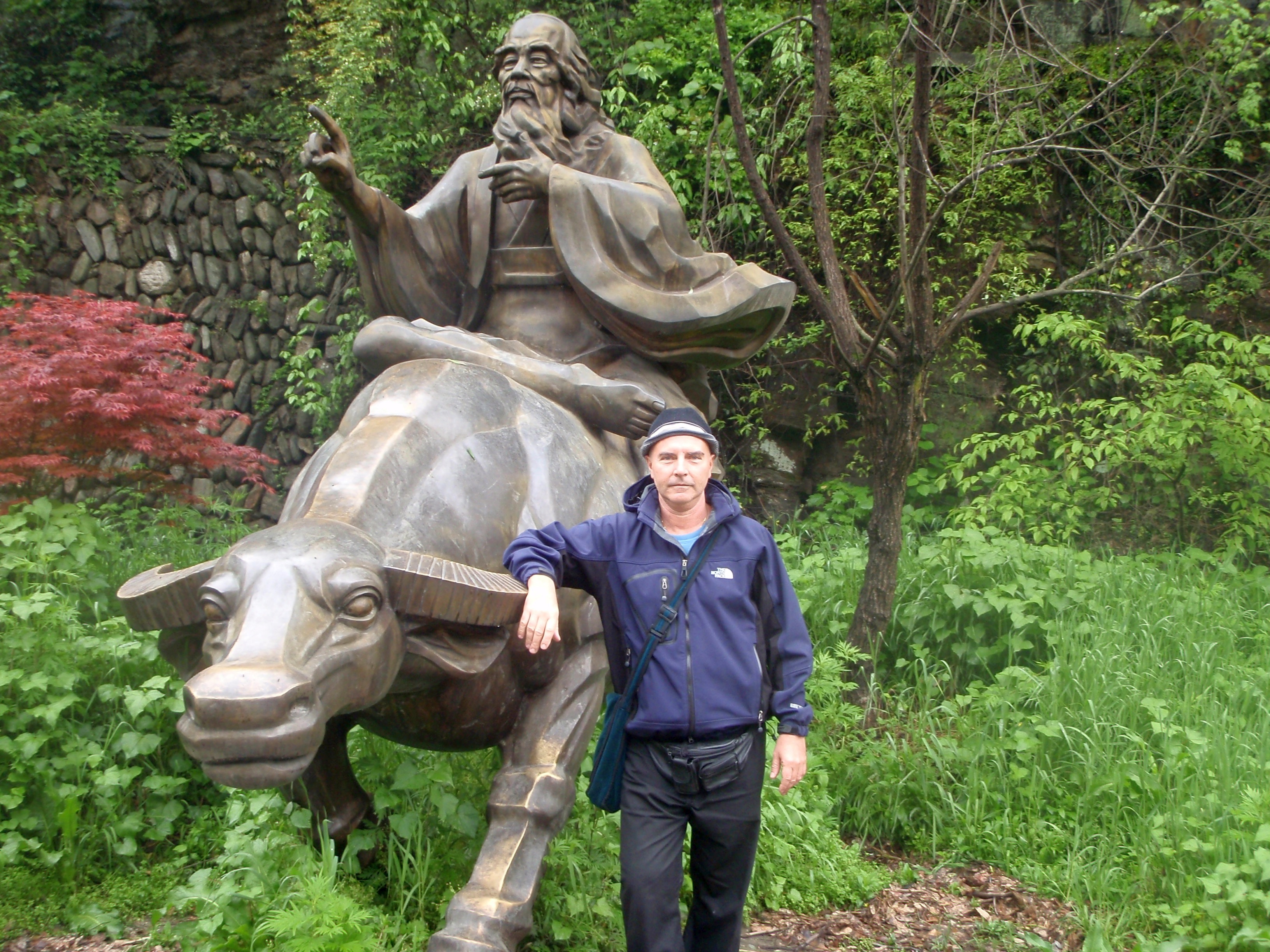 Kadama beside a large bronze statue of Lao Tzu riding a water buffalo, green forest background