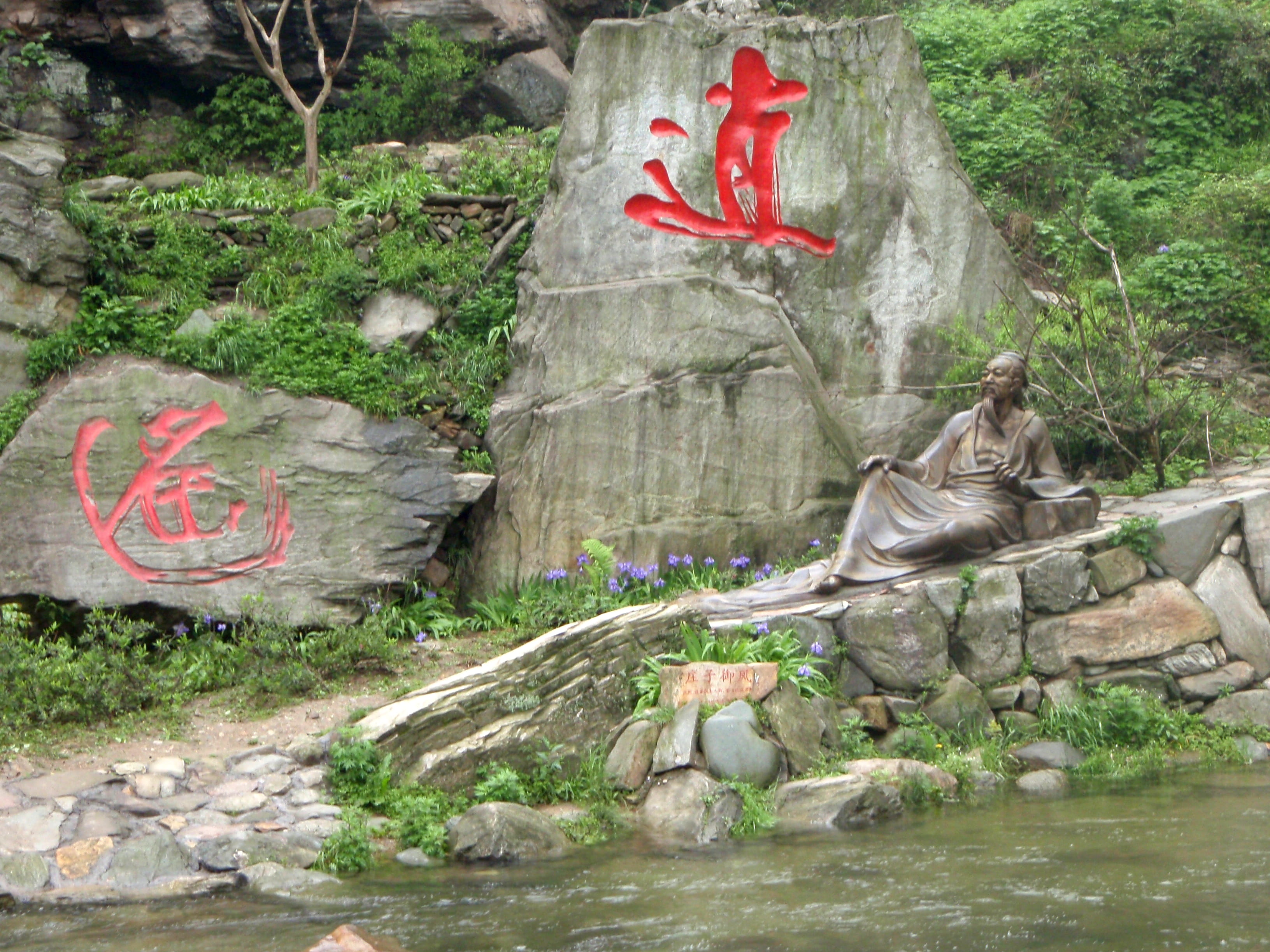 Serene rock garden with bronze Taoist sage statue seated by a stream, red calligraphy on stone, lush greenery