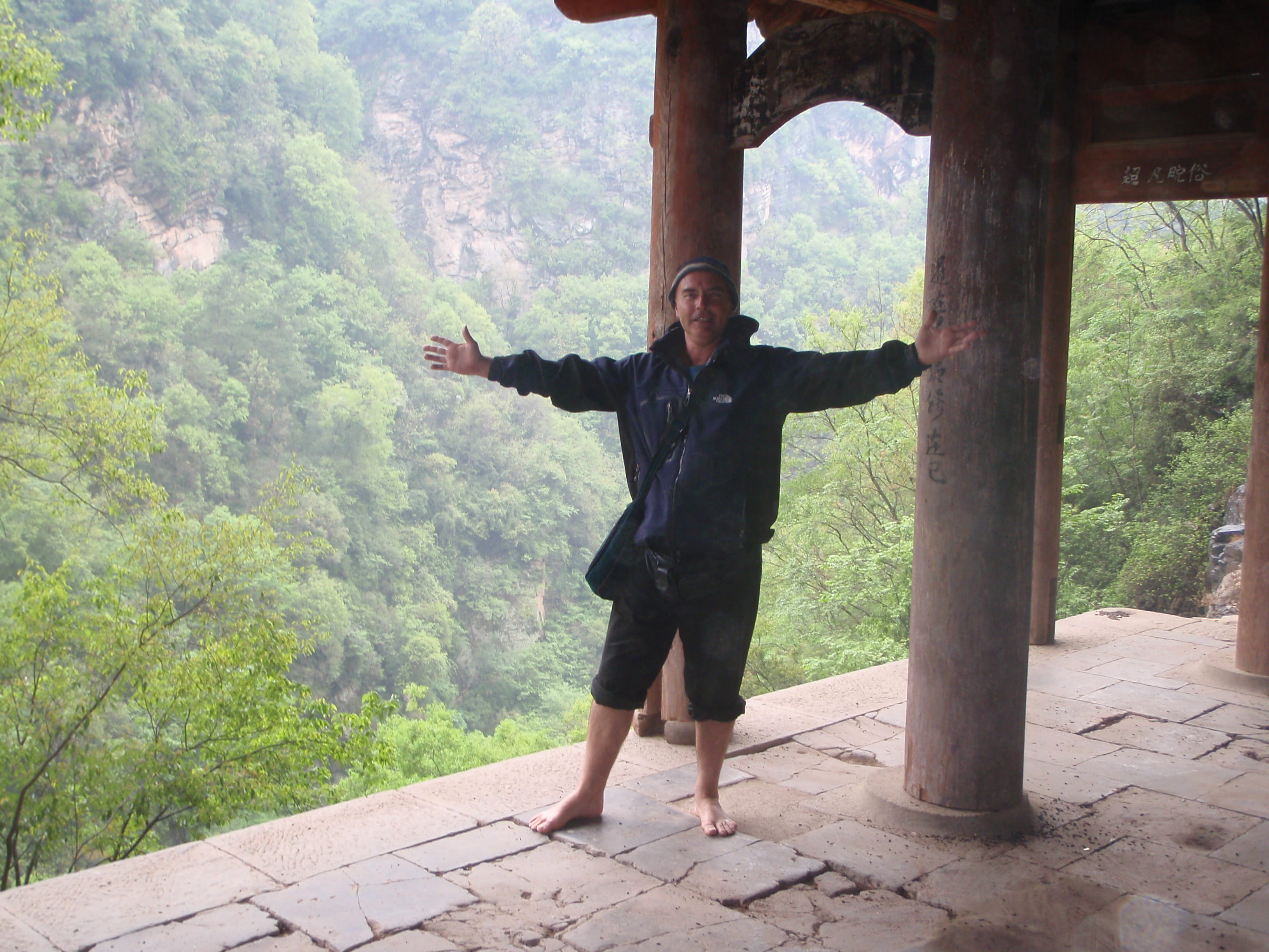 Kadama in casual black jacket with arms outstretched wide in welcoming gesture, barefoot in misty temple pavilion with green valley behind