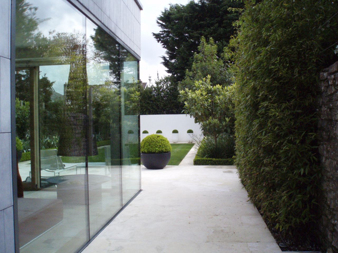 Sleek limestone terrace beside a glass-walled house with a tall hedge and clipped box sphere