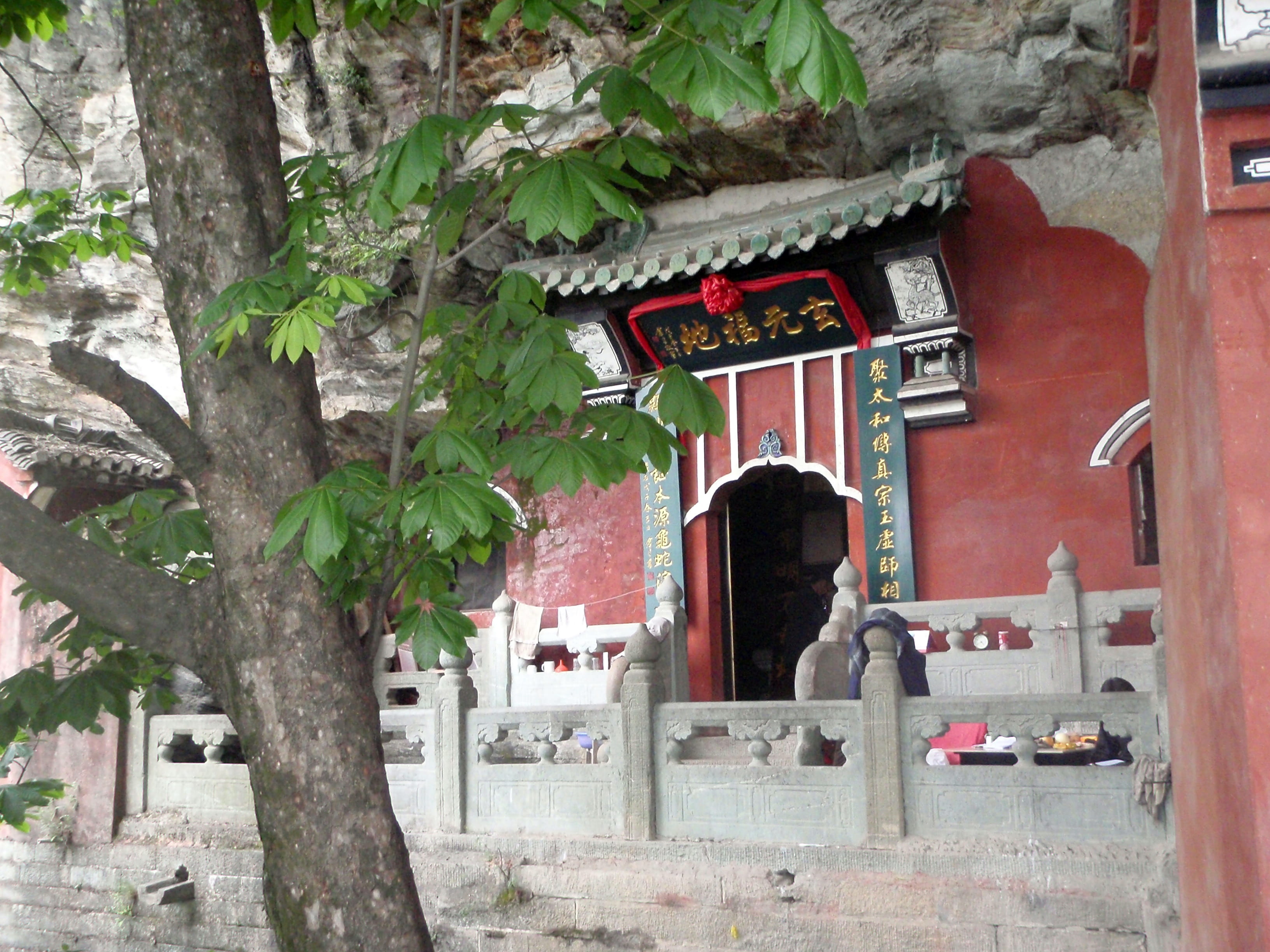 Red-lacquered temple entrance with traditional carved pillars, green trees framing, quiet natural setting