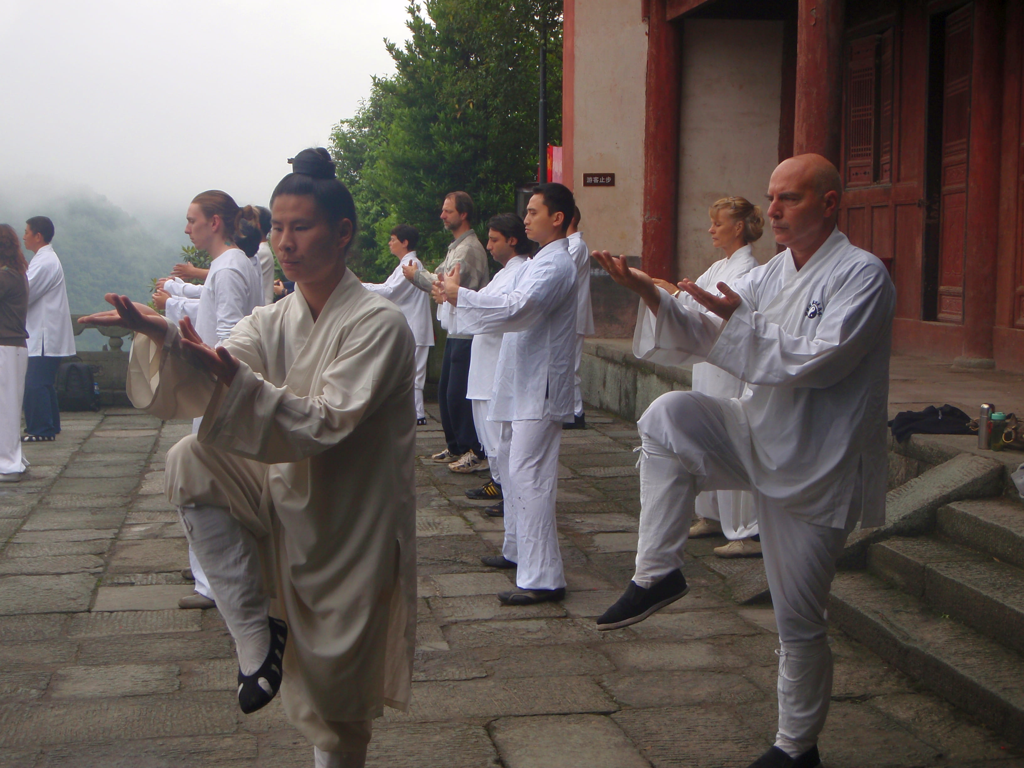 Misty morning terrace practice, students in white and beige robes performing synchronized single-leg stance