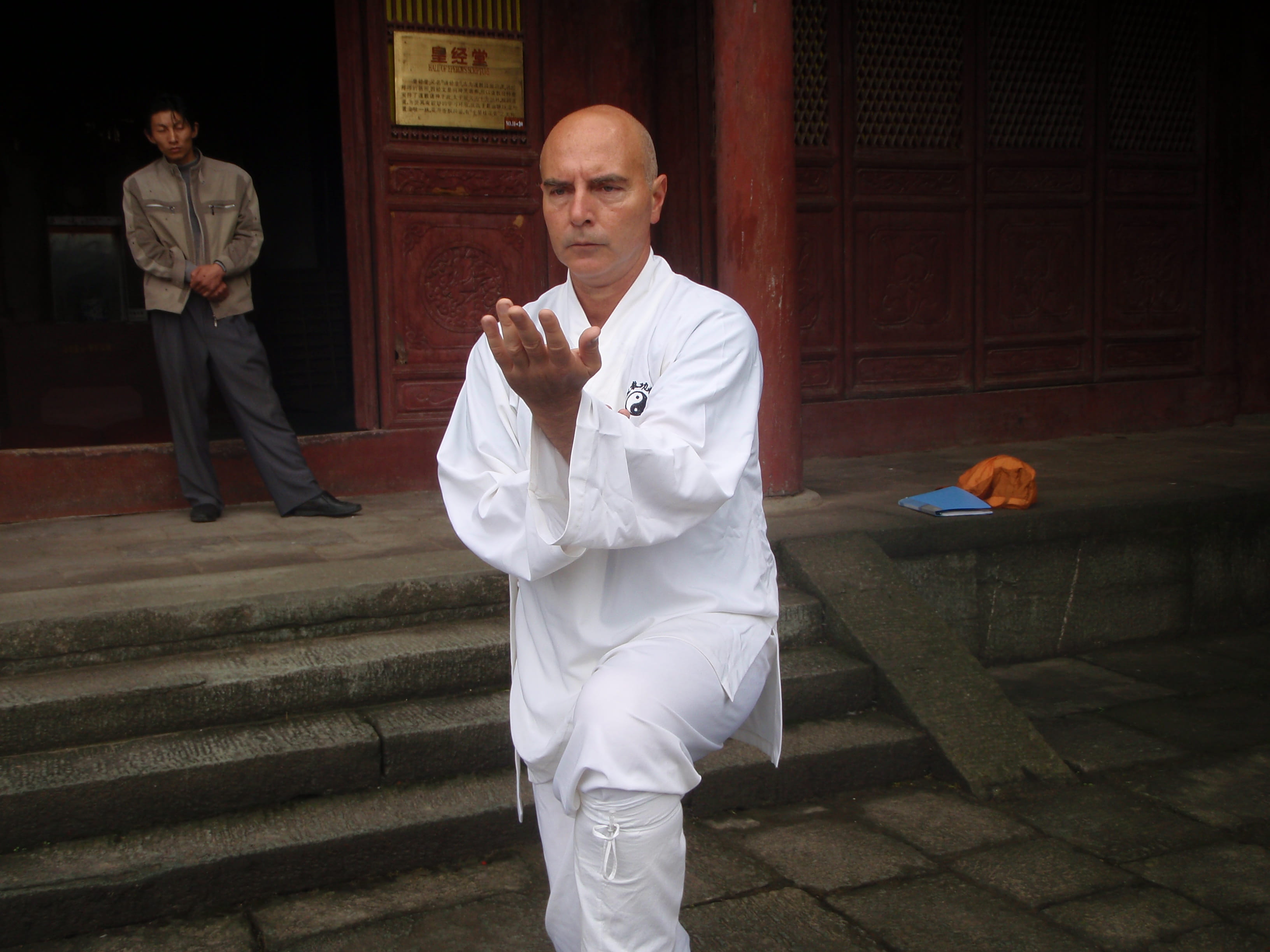 Kadama demonstrating a qigong hand position on dark temple steps, student in traditional dress observing at left