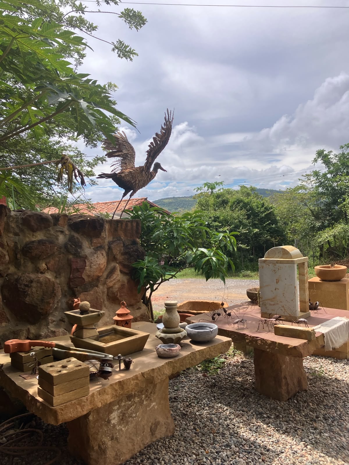 Escultura de una garza con alas abiertas junto a figuras de hormigas y piezas de cerámica sobre mesas de piedra en el exterior