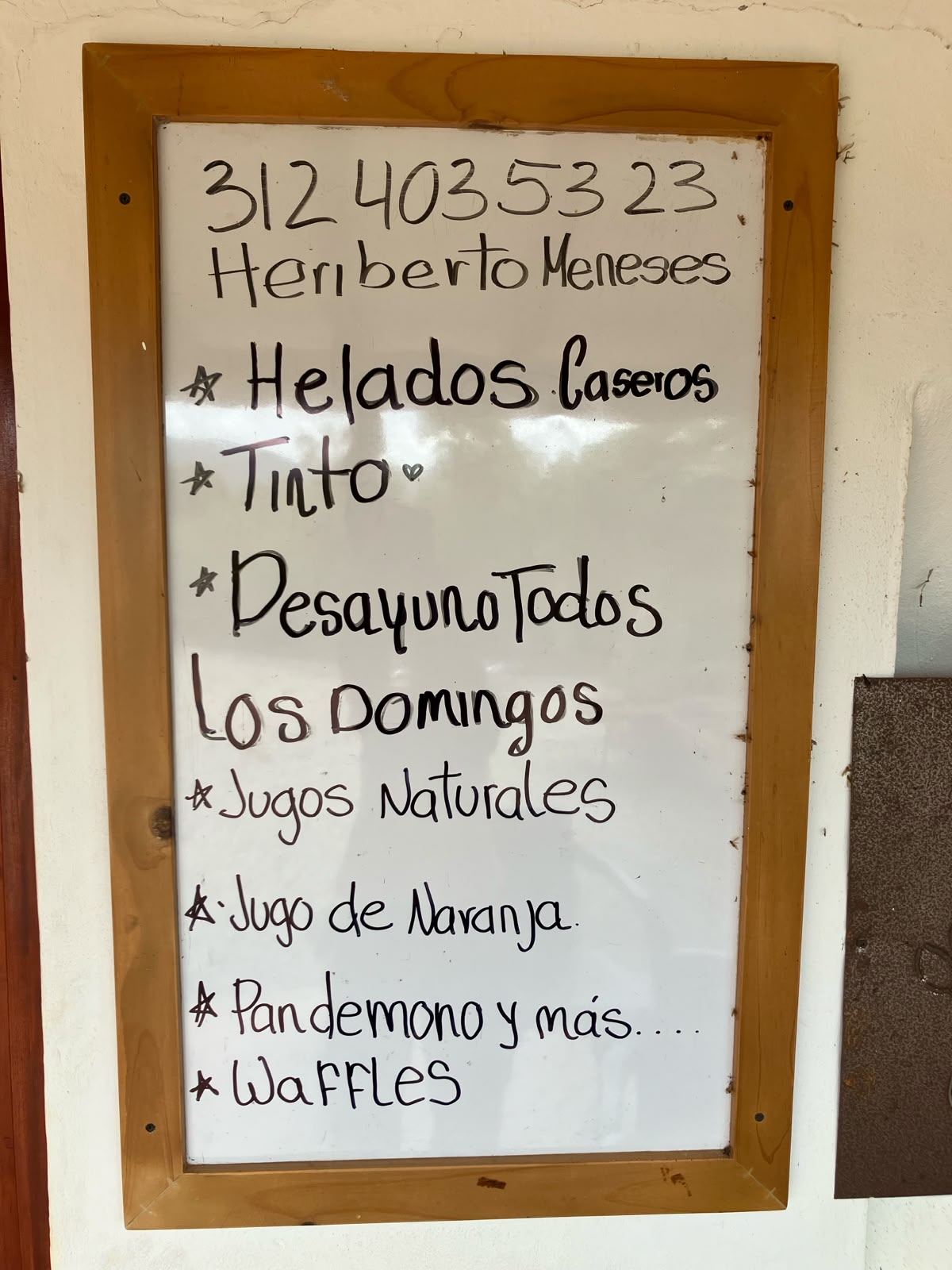 Tablero blanco con marco de madera que muestra el menú escrito a mano: helados caseros, tinto, desayuno todos los domingos, jugos naturales, pandebono y waffles