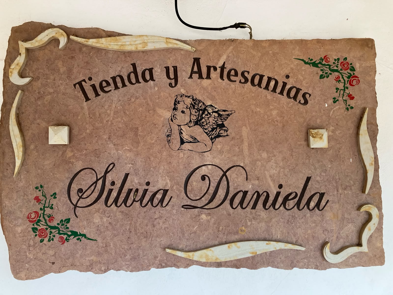 Letrero en piedra con la inscripción Tienda y Artesanías Silvia Daniela con un ángel grabado y ramas de rosas decorativas