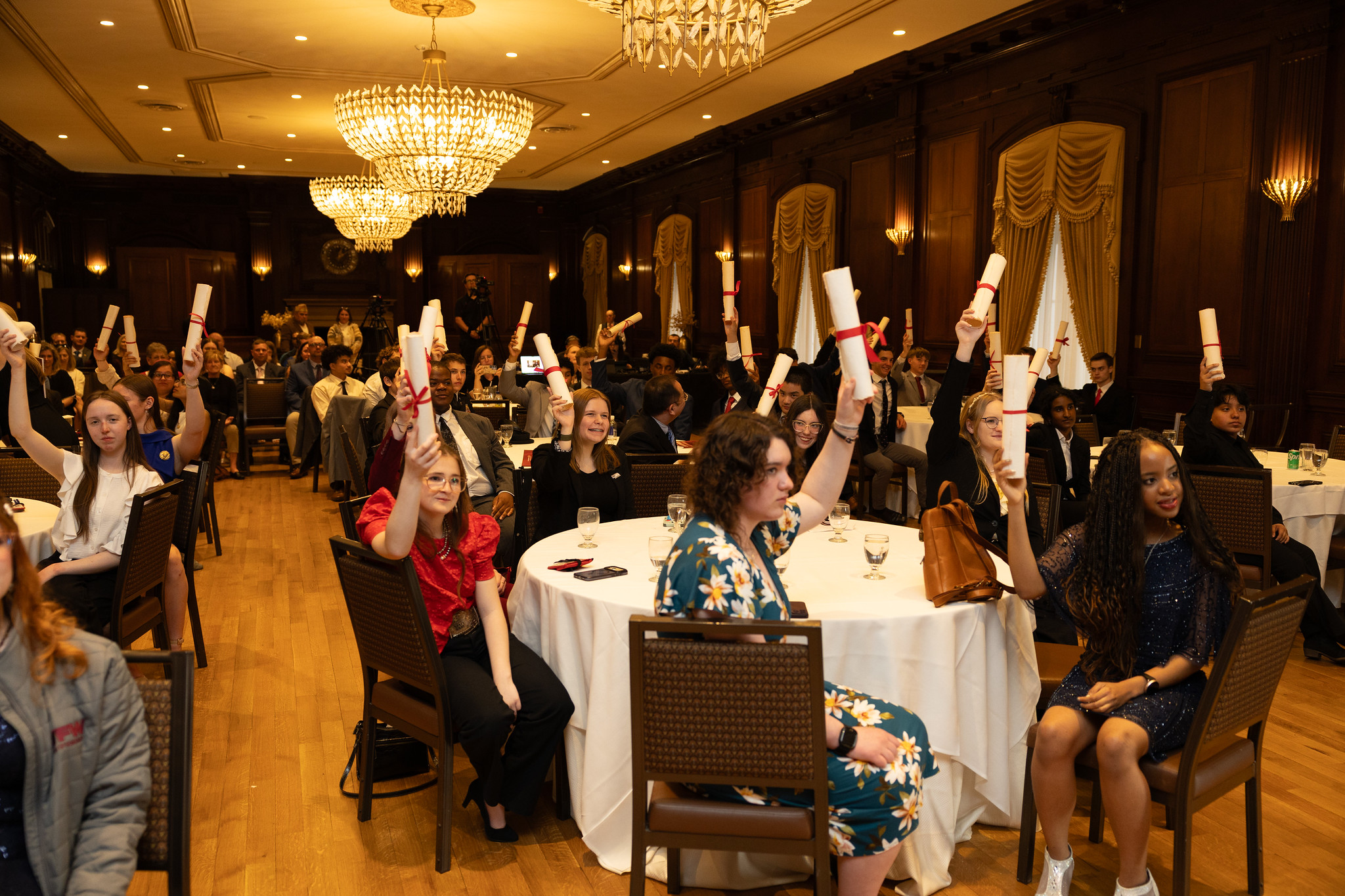 Patriot's Pen student winners raising rolled award certificates tied with red ribbons at a formal recognition banquet