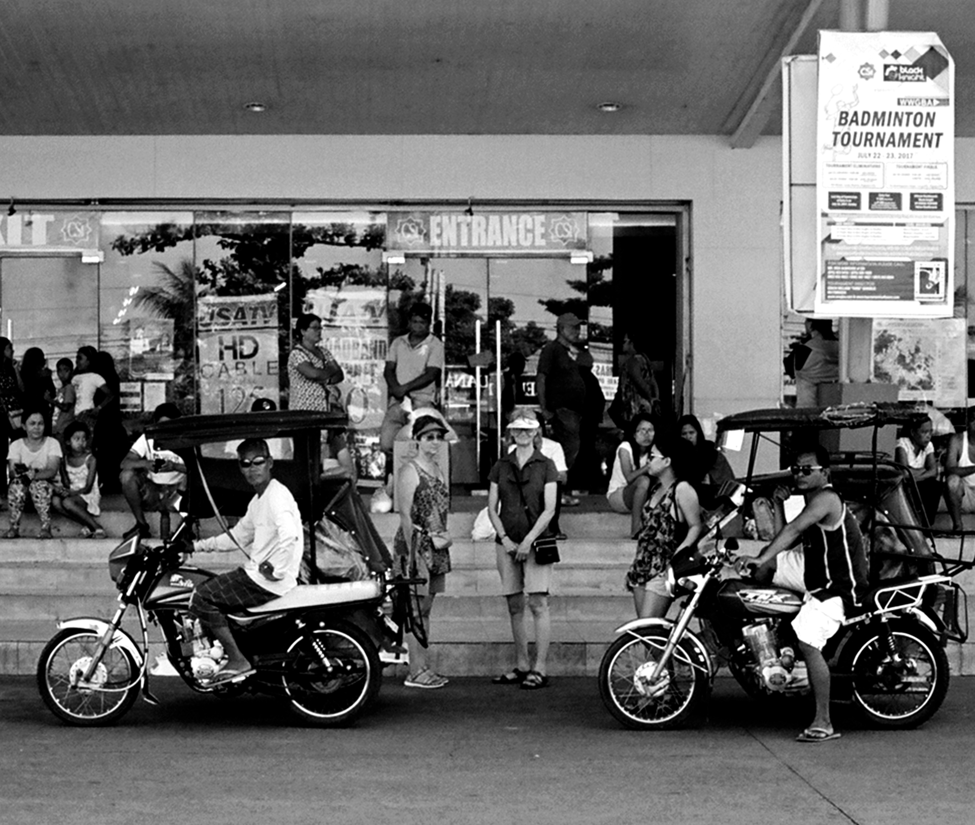 Urban street scene with motorcycles and people near shopping area with badminton tournament sign