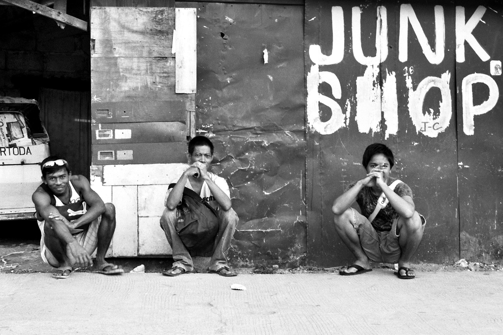 Three men squatting against wall with Junk Shop sign painted behind them