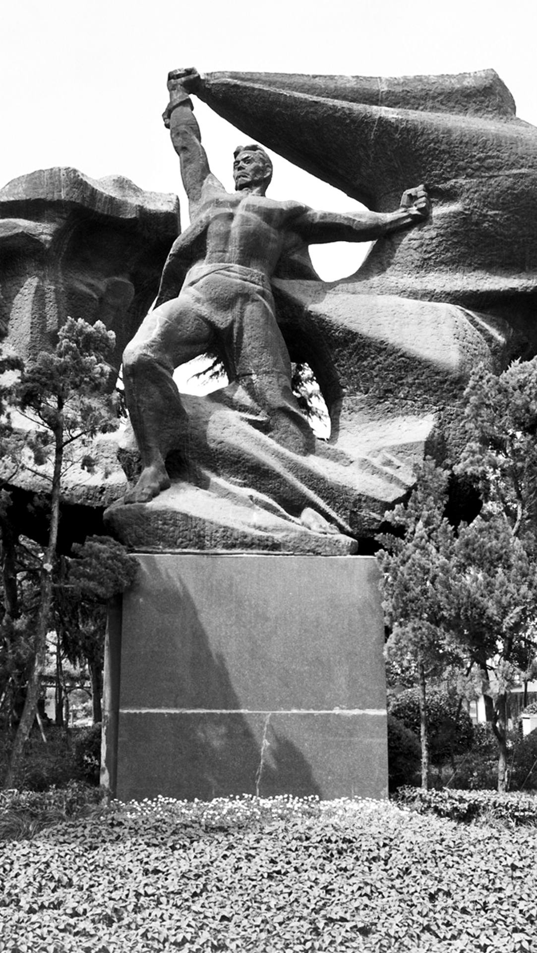 Large stone statue of liberation figure with arms raised against sky and foliage