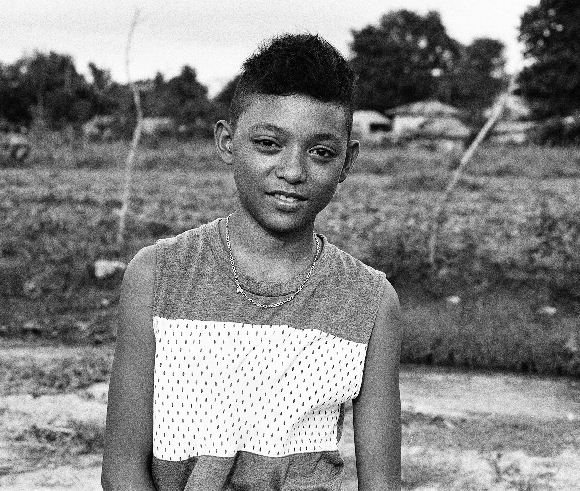 Black and white portrait of young boy with animated smile wearing patterned shirt in rural setting