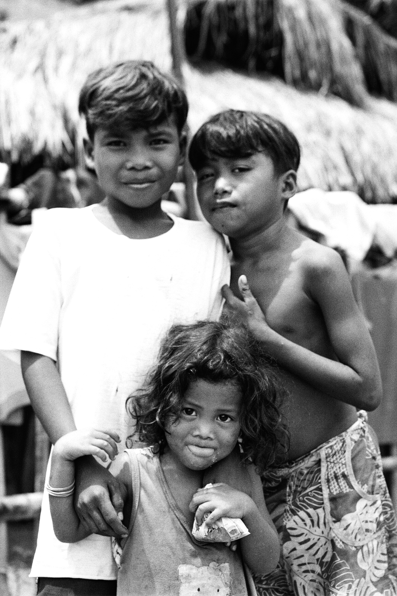 Black and white portrait of three children standing together outdoors with thatched roof background