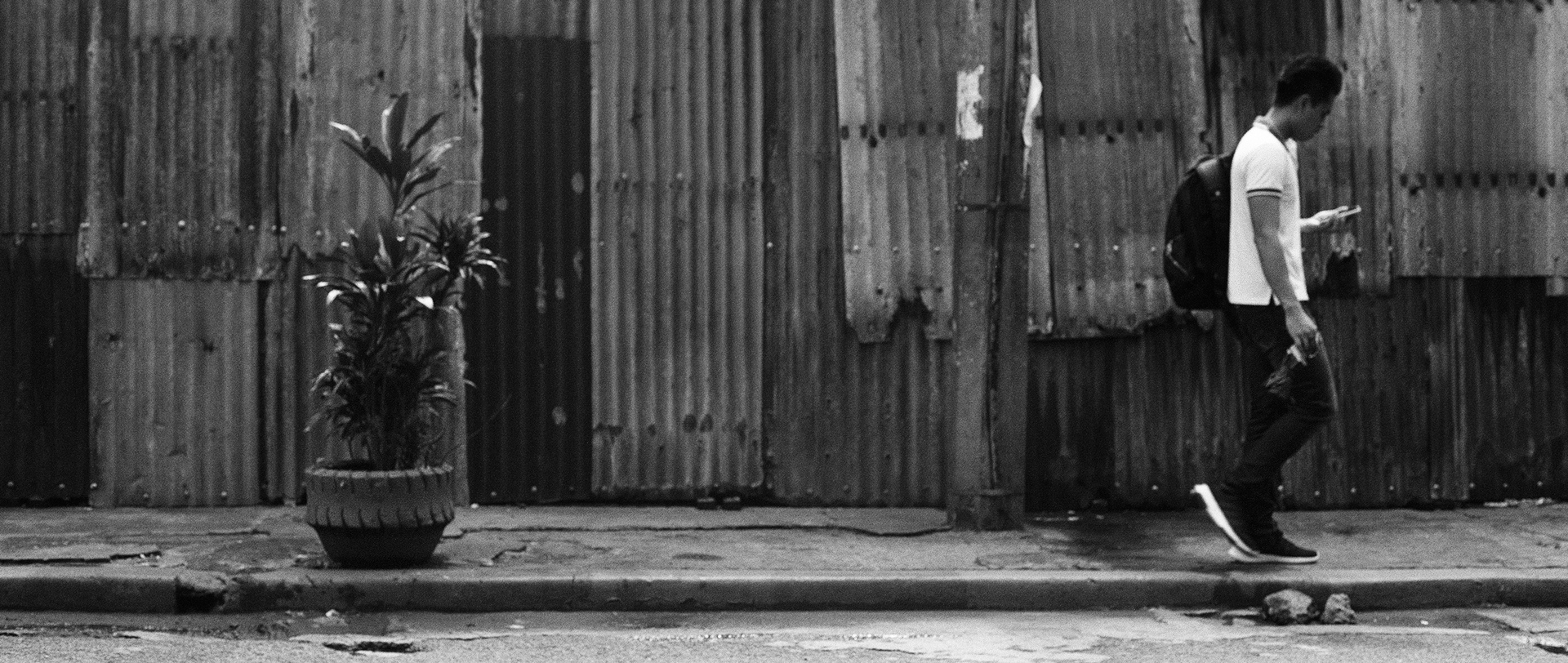 Black and white man in tank top walking past weathered corrugated metal wall with potted plant