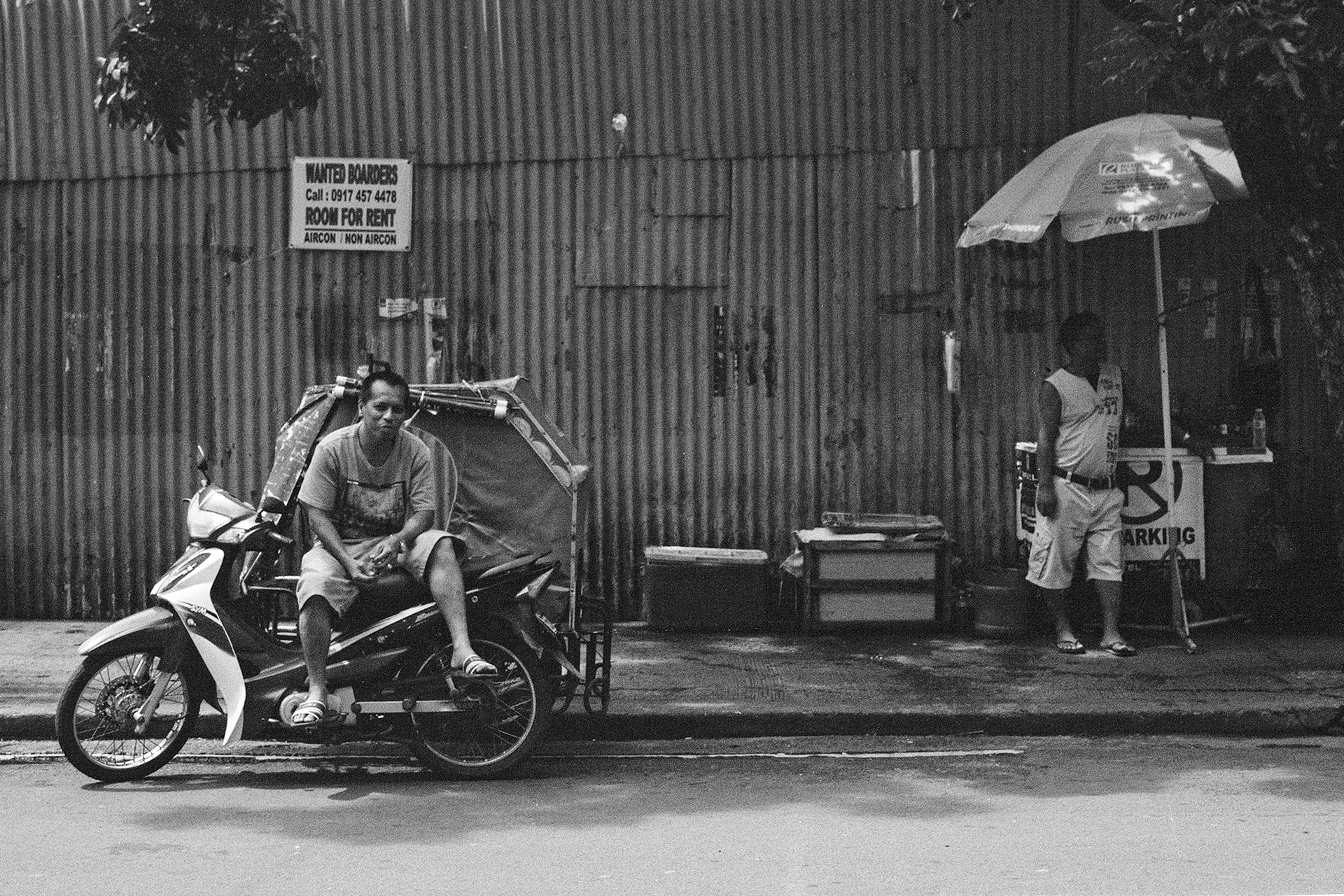 Black and white man sitting on motorcycle sidecar against corrugated metal building with signage