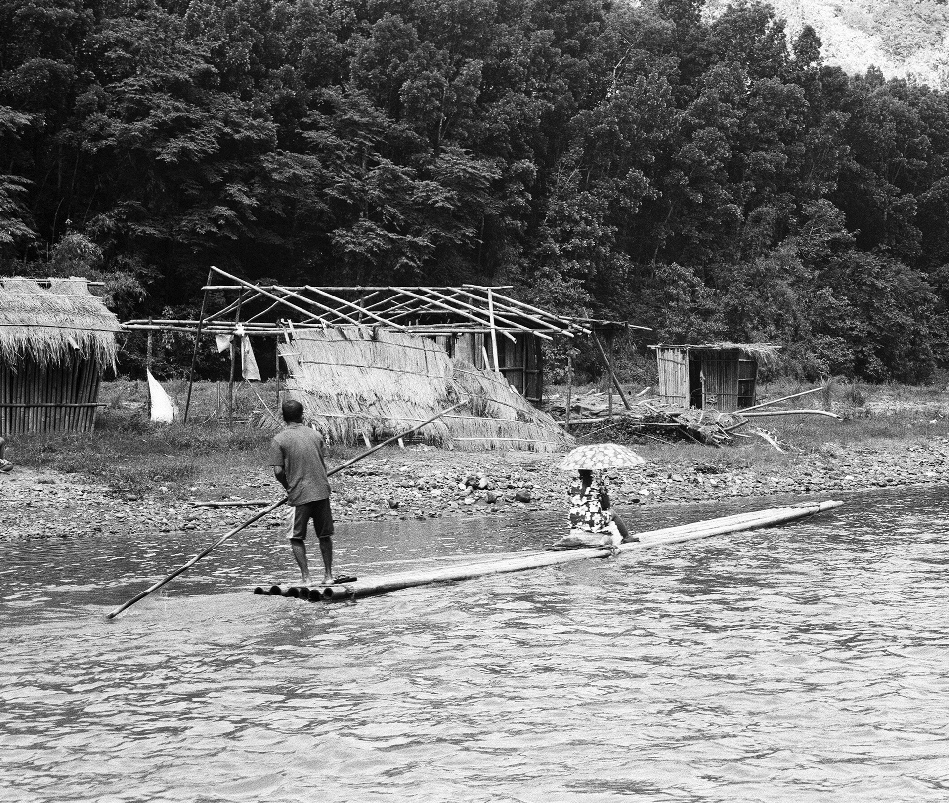 Black and white man poling log raft in river with thatched structures and forested hillside background
