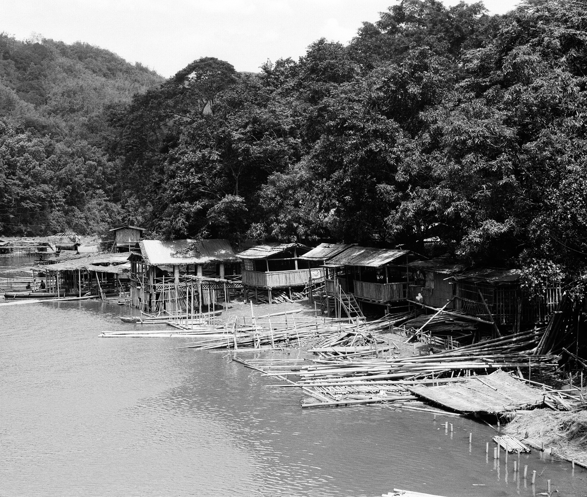 Black and white waterfront settlement wooden stilt structures reflected in calm water forested hills