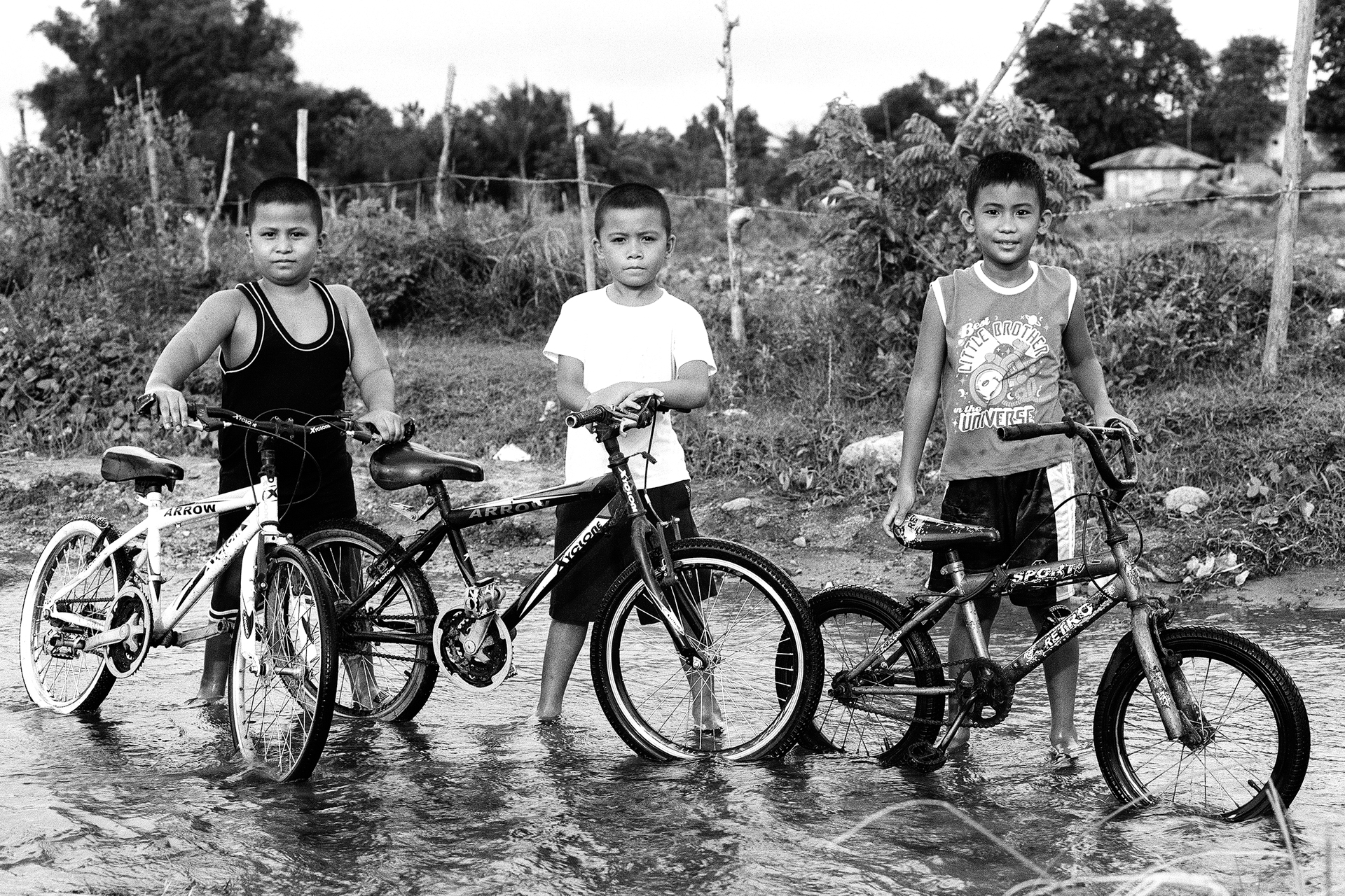 Black and white three young boys with bicycles standing in flooded water area rural setting