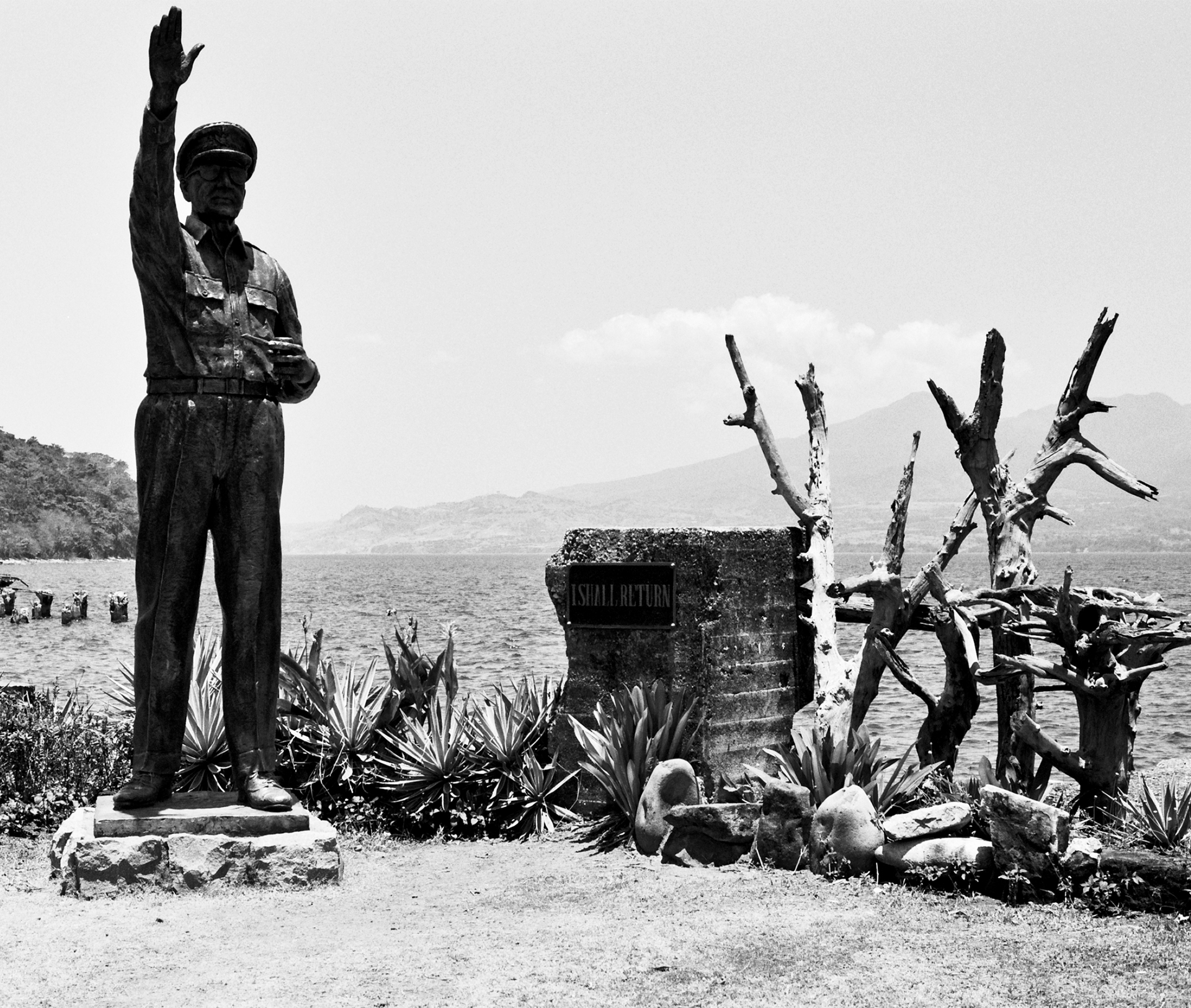 Black and white memorial statue with arms raised overlooking water with dead trees and church