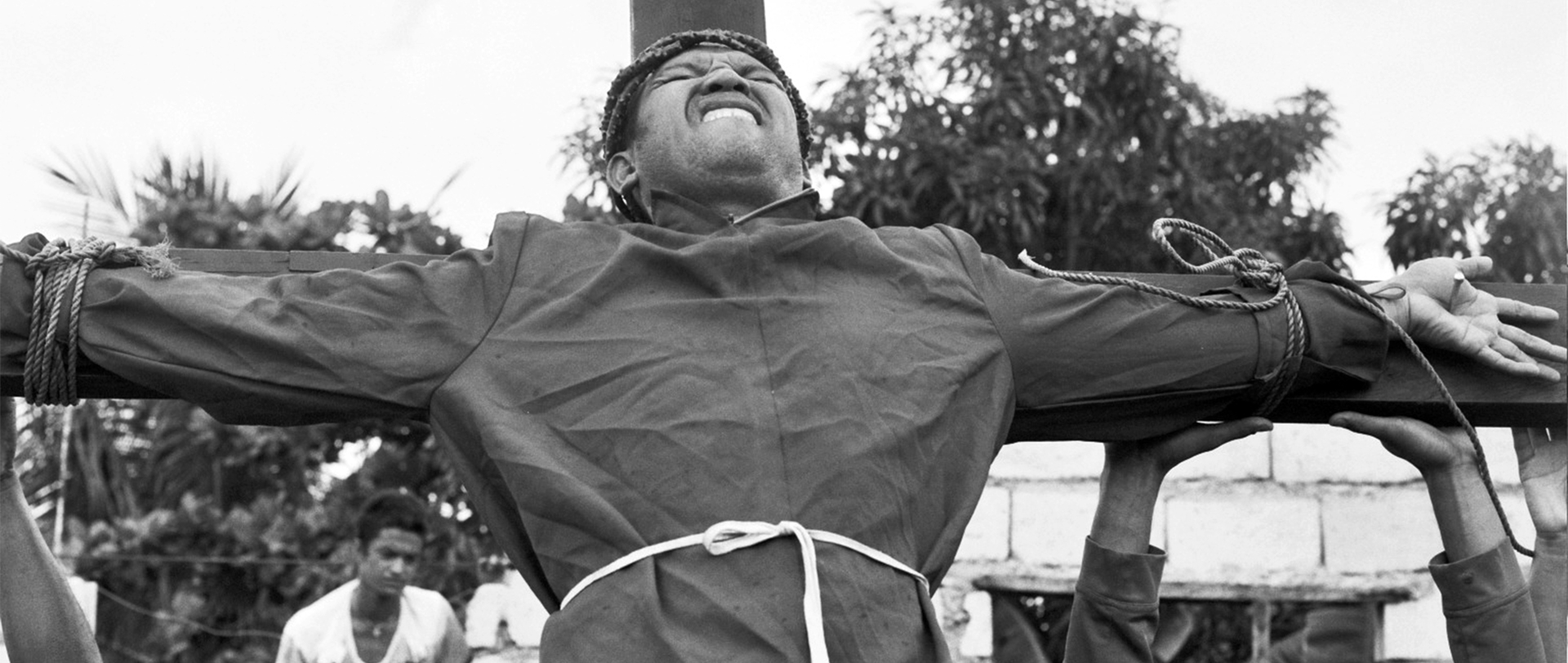 Black and white man in crucifixion pose against wooden cross with rope bindings outdoors