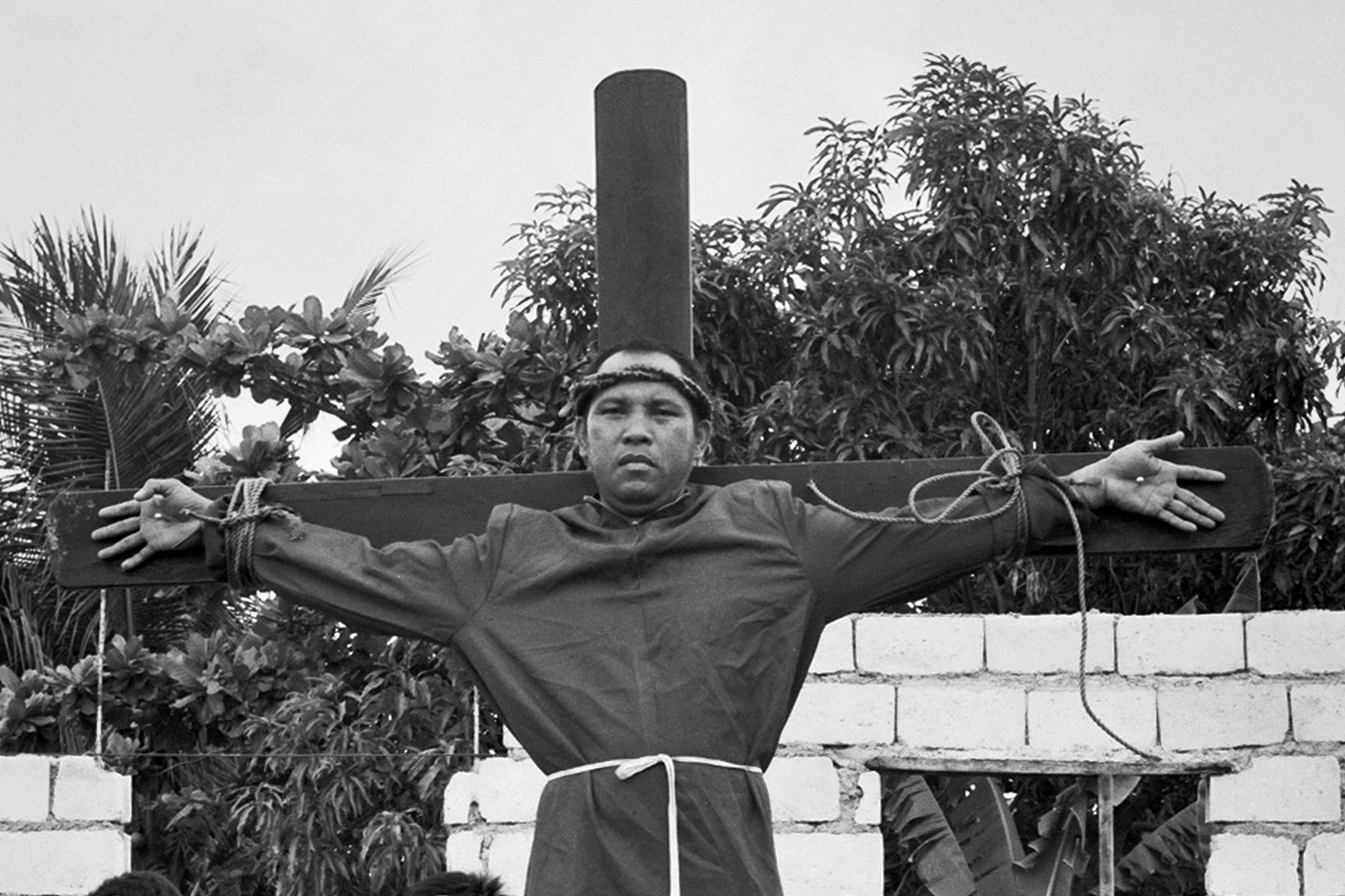 Black and white man with arms extended horizontally against wooden cross in garden setting