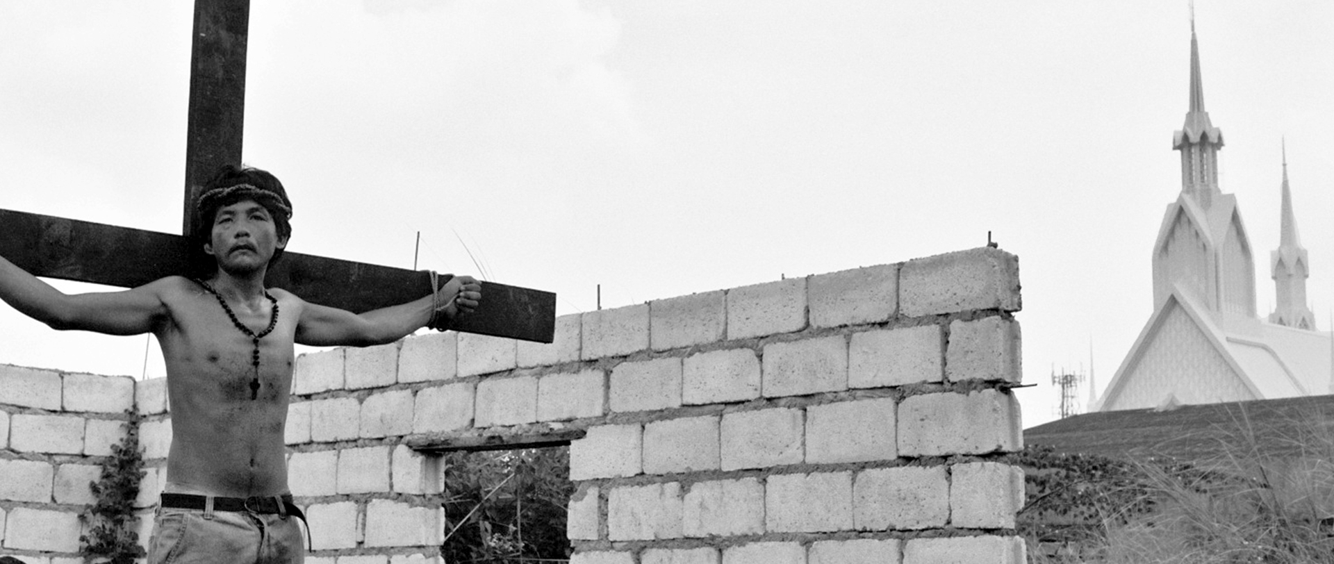 Black and white man with arms outstretched against large wooden cross with church backdrop