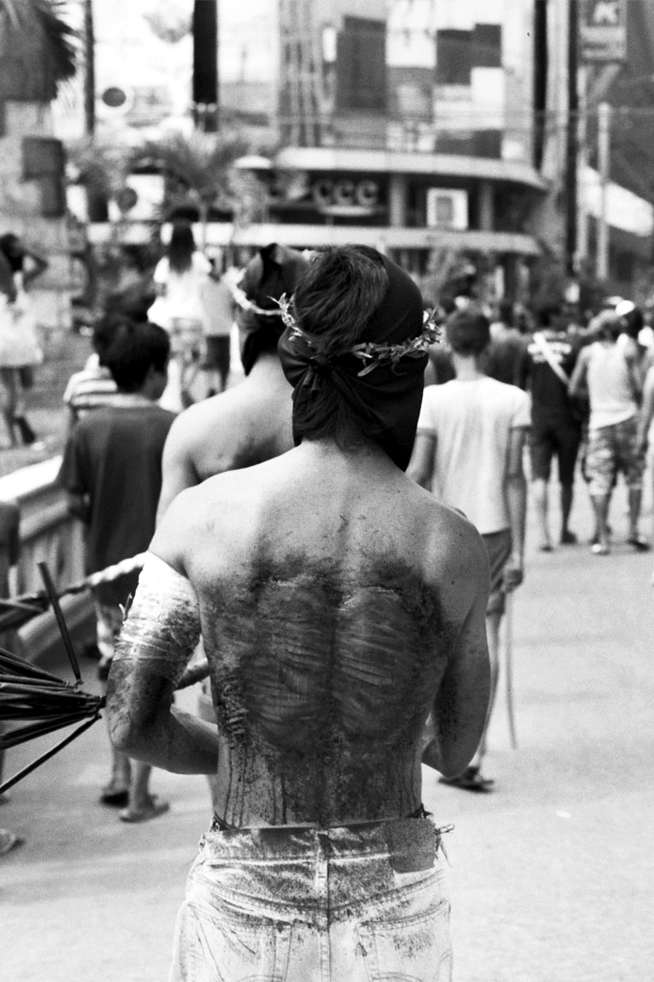 Black and white figure from behind wearing flower wreath in busy street scene with crowds