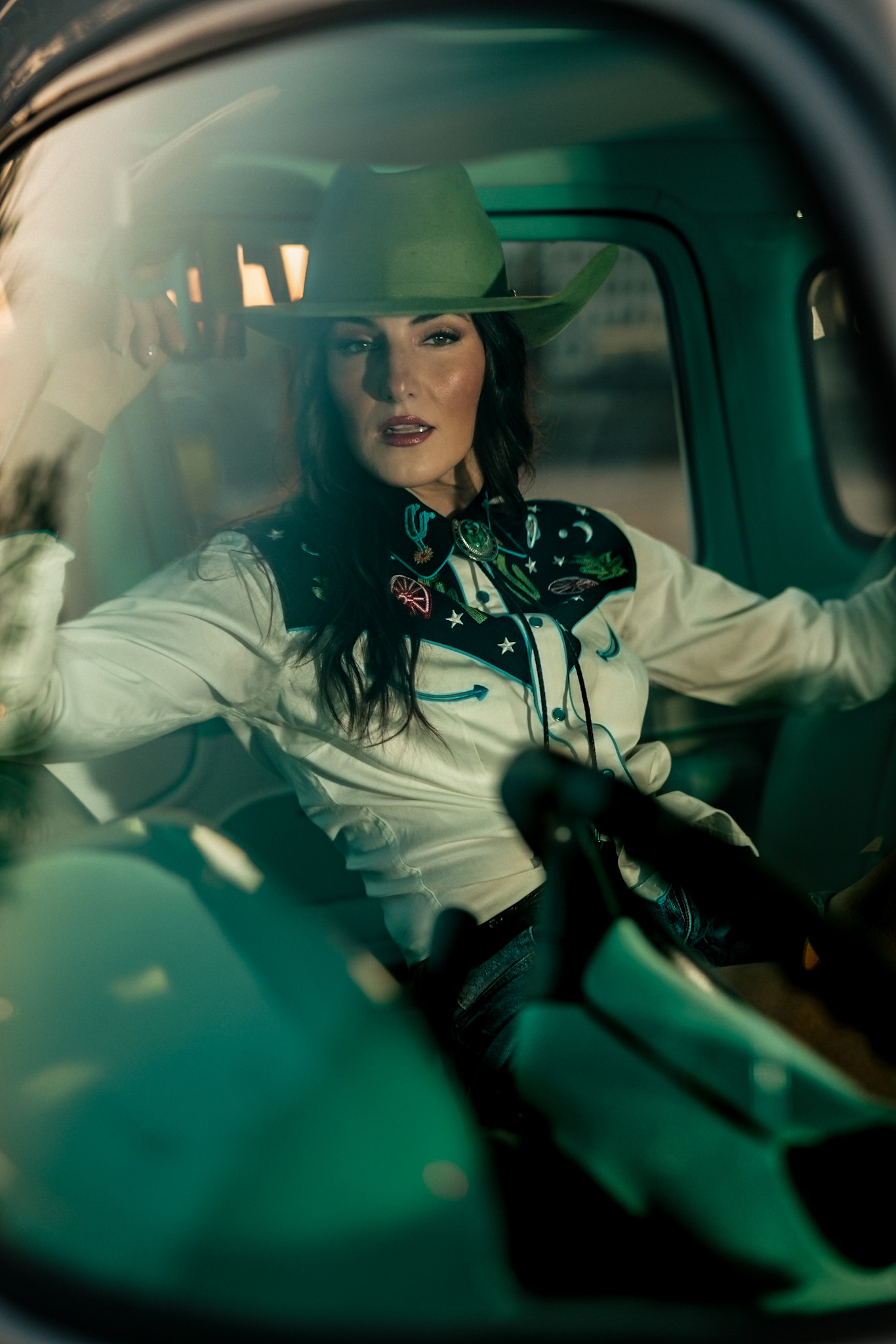 Model in a green cowboy hat and embroidered Western shirt behind the wheel of a vintage truck, lit in cinematic teal