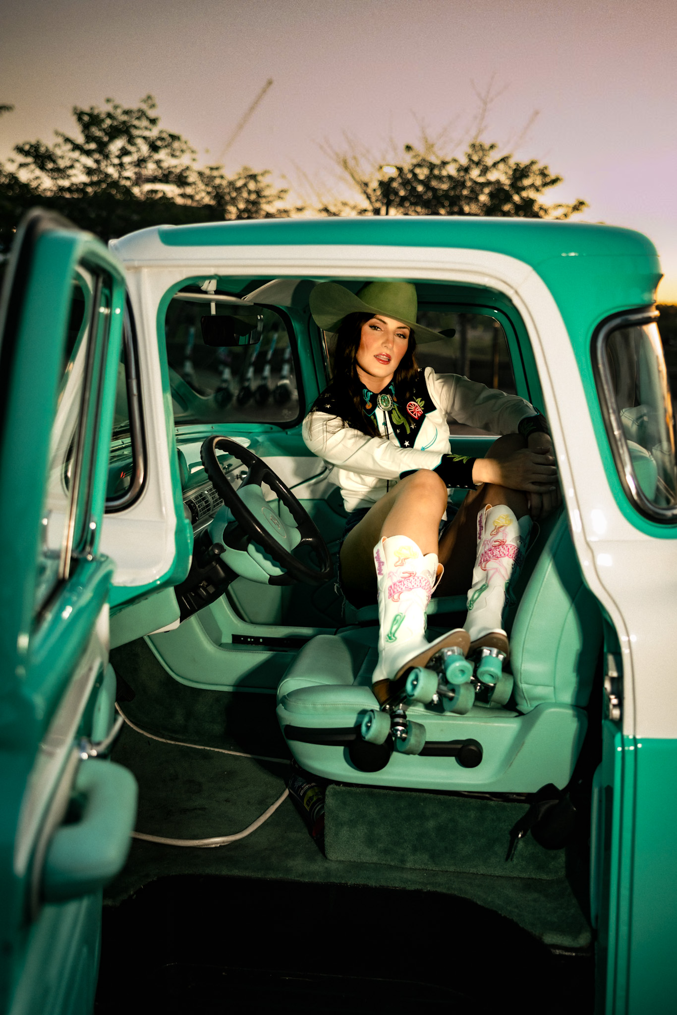 Model in a green hat resting inside a teal vintage truck with white roller-skate cowboy boots propped up
