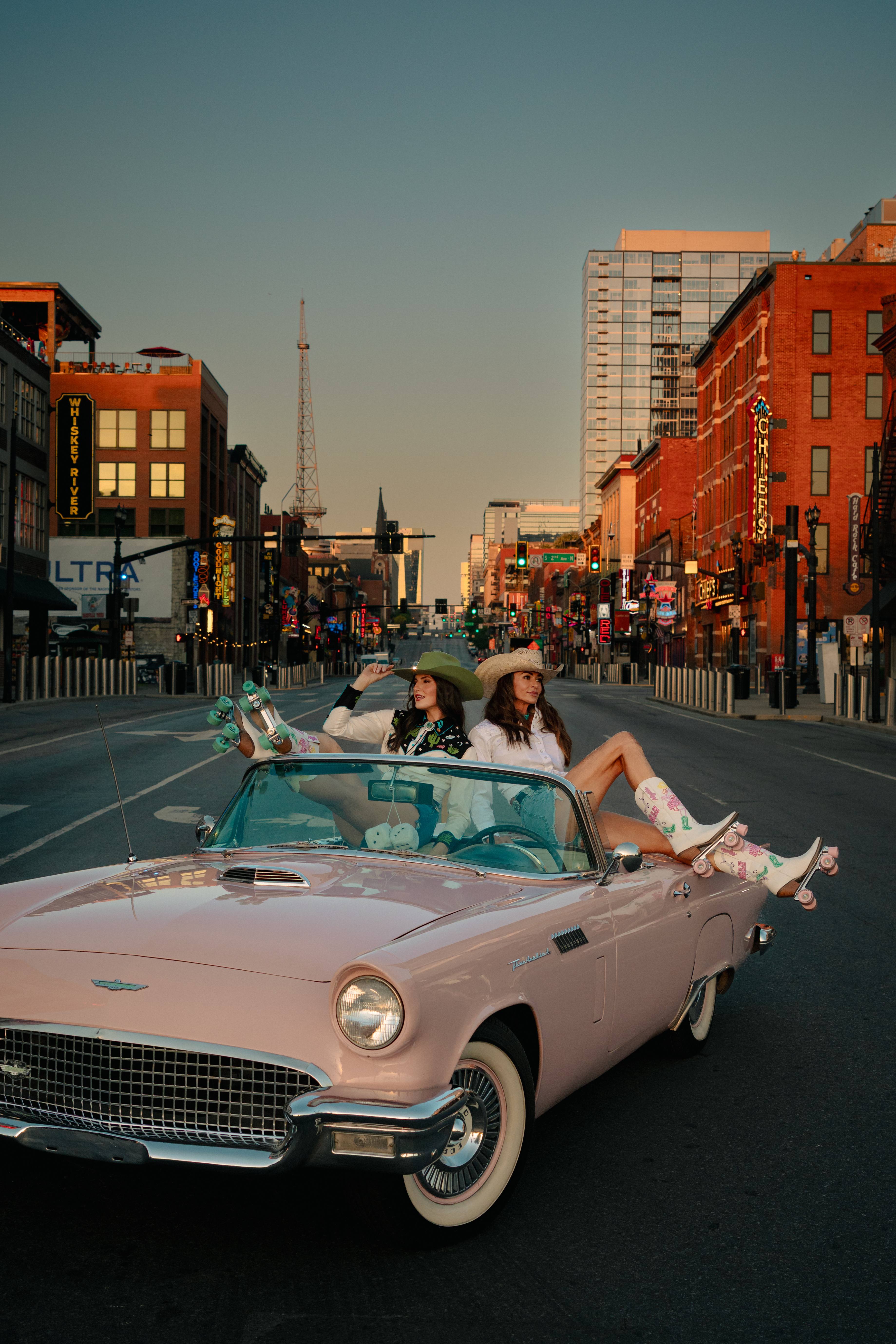 Two women in Western wear and roller-skate boots lounging on a pink vintage convertible on Broadway in downtown Nashville