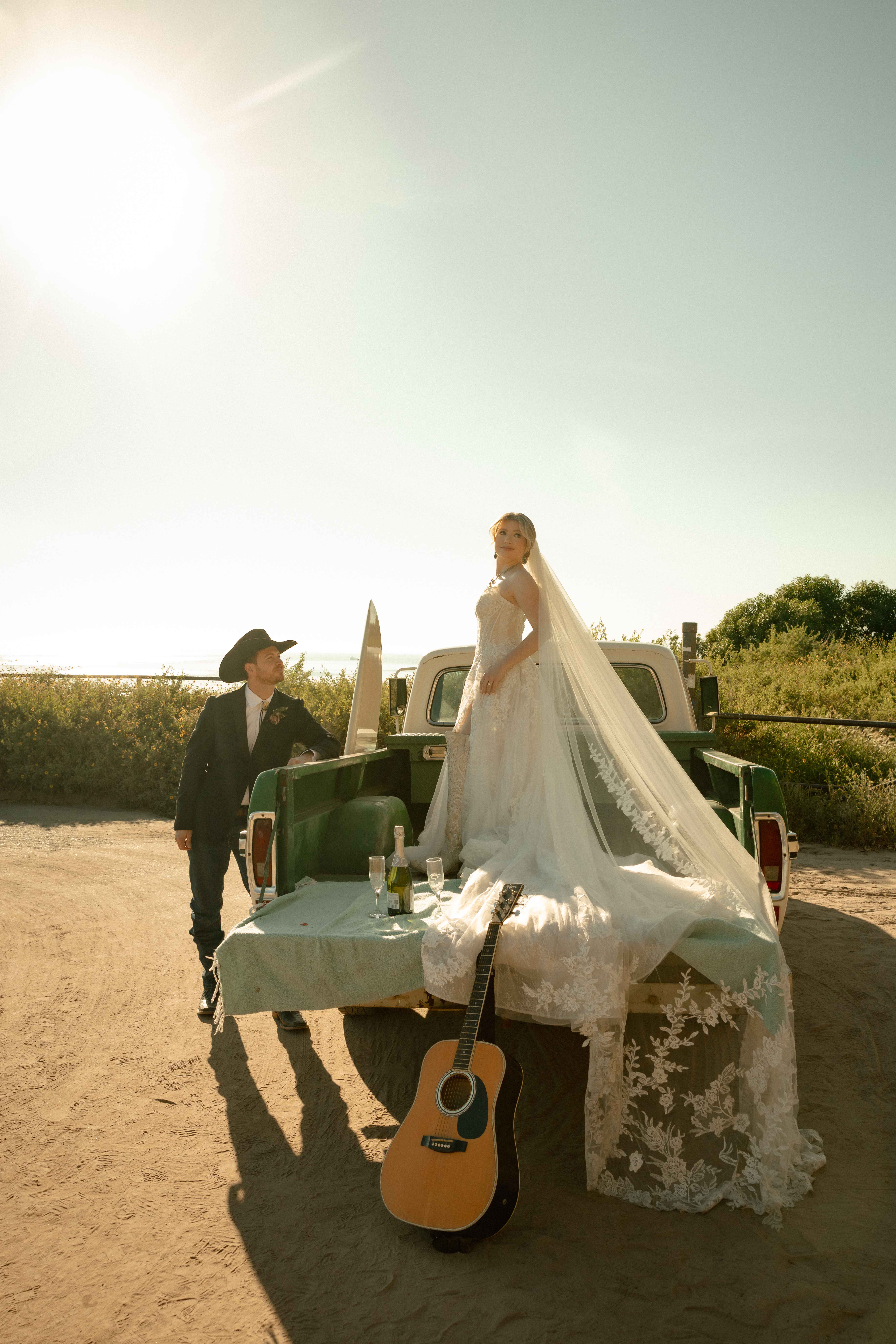 Bride in a lace gown standing on the tailgate of a vintage green pickup truck at golden hour, with a groom in a cowboy hat and an acoustic guitar resting against the truck