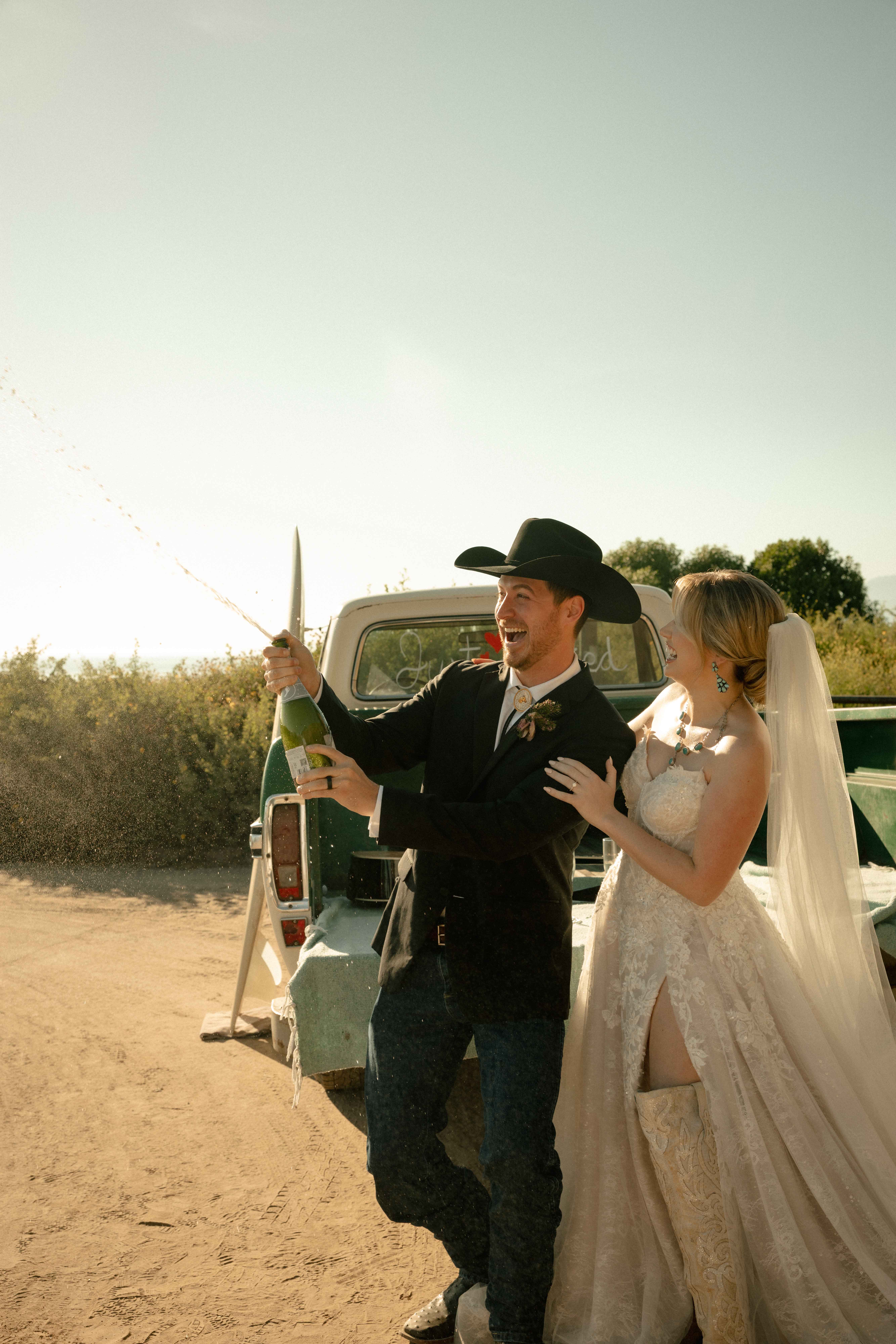 Groom in a black cowboy hat spraying champagne beside a laughing bride at a vintage truck in golden light