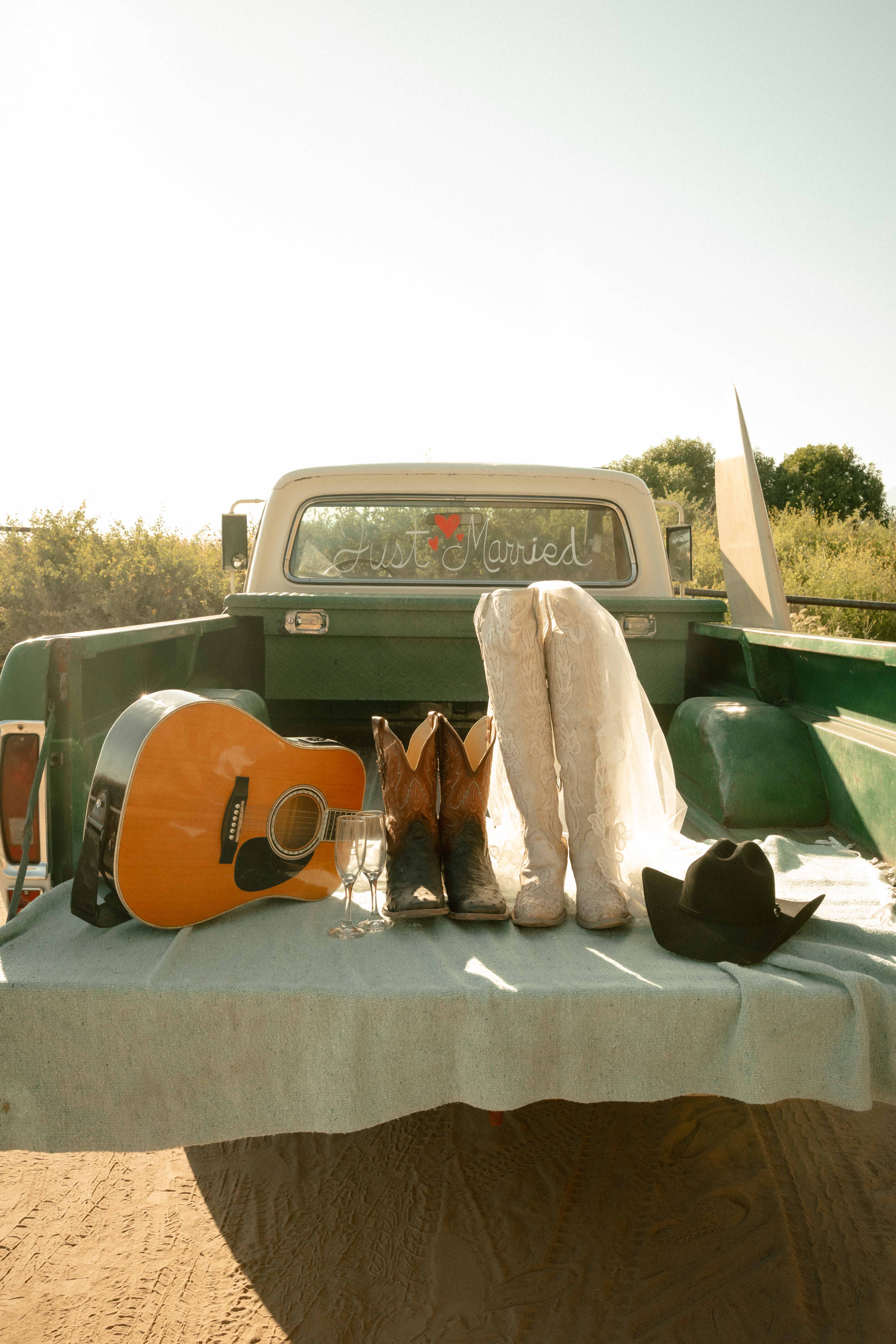 Editorial detail of cowboy boots, a lace gown, a black hat and an acoustic guitar arranged on a truck bed with a Just Married window sign