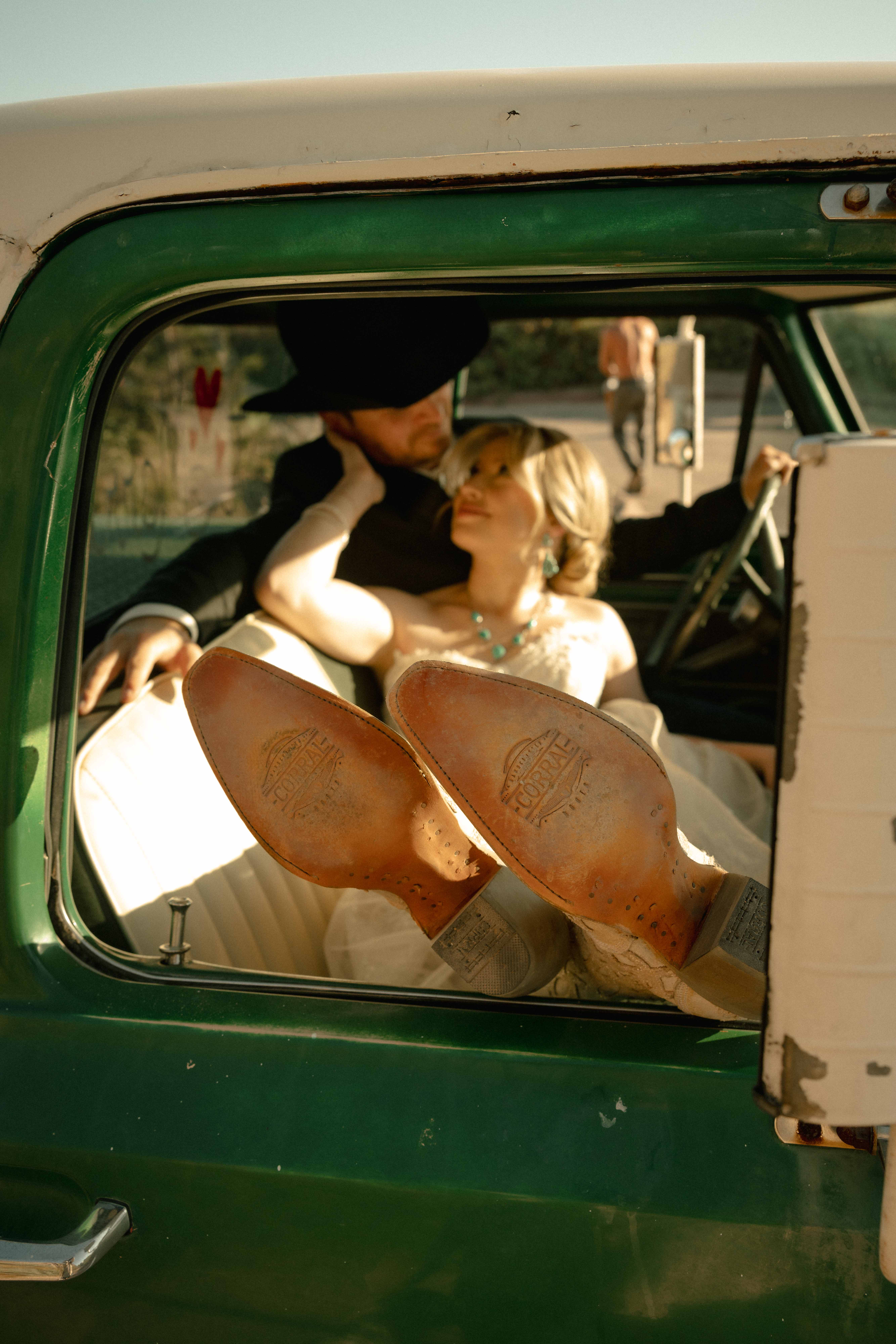 Close-up of Corral-branded leather boot soles resting in a vintage green truck window as a couple embraces inside
