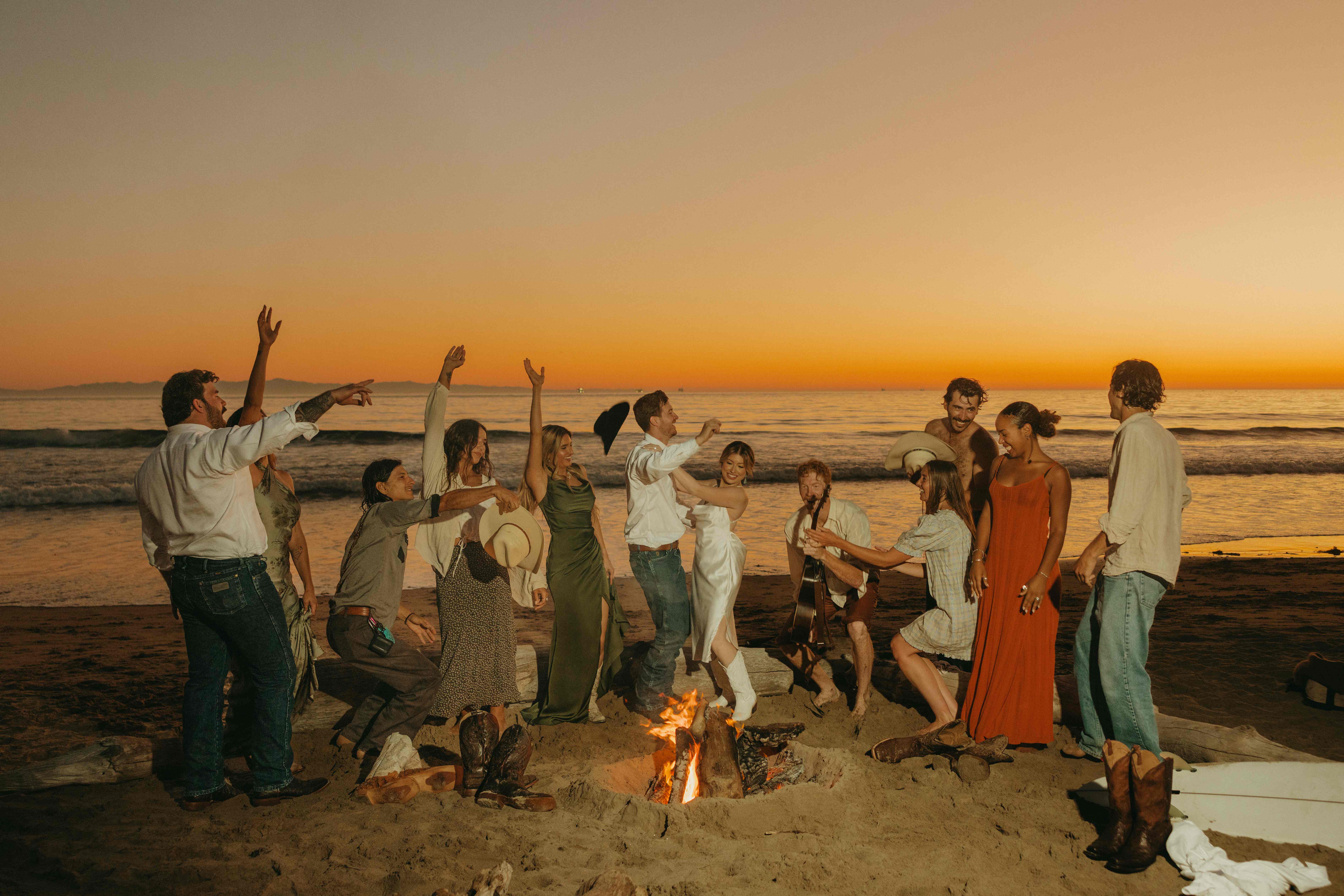 Group of friends dancing and celebrating around a beach bonfire at sunset
