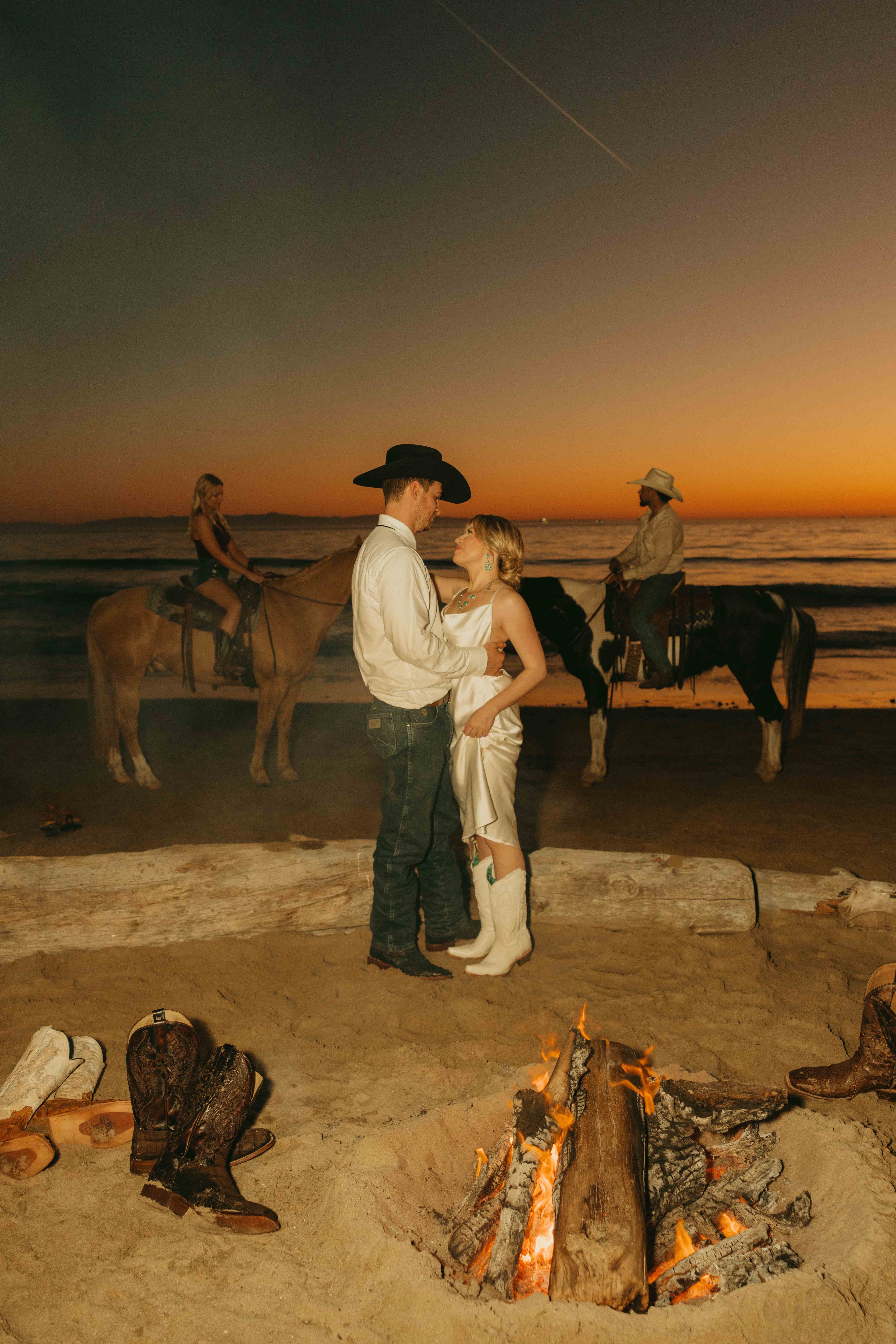 Couple in Western wedding attire embracing beside a beach bonfire at dusk while two riders wait on horseback