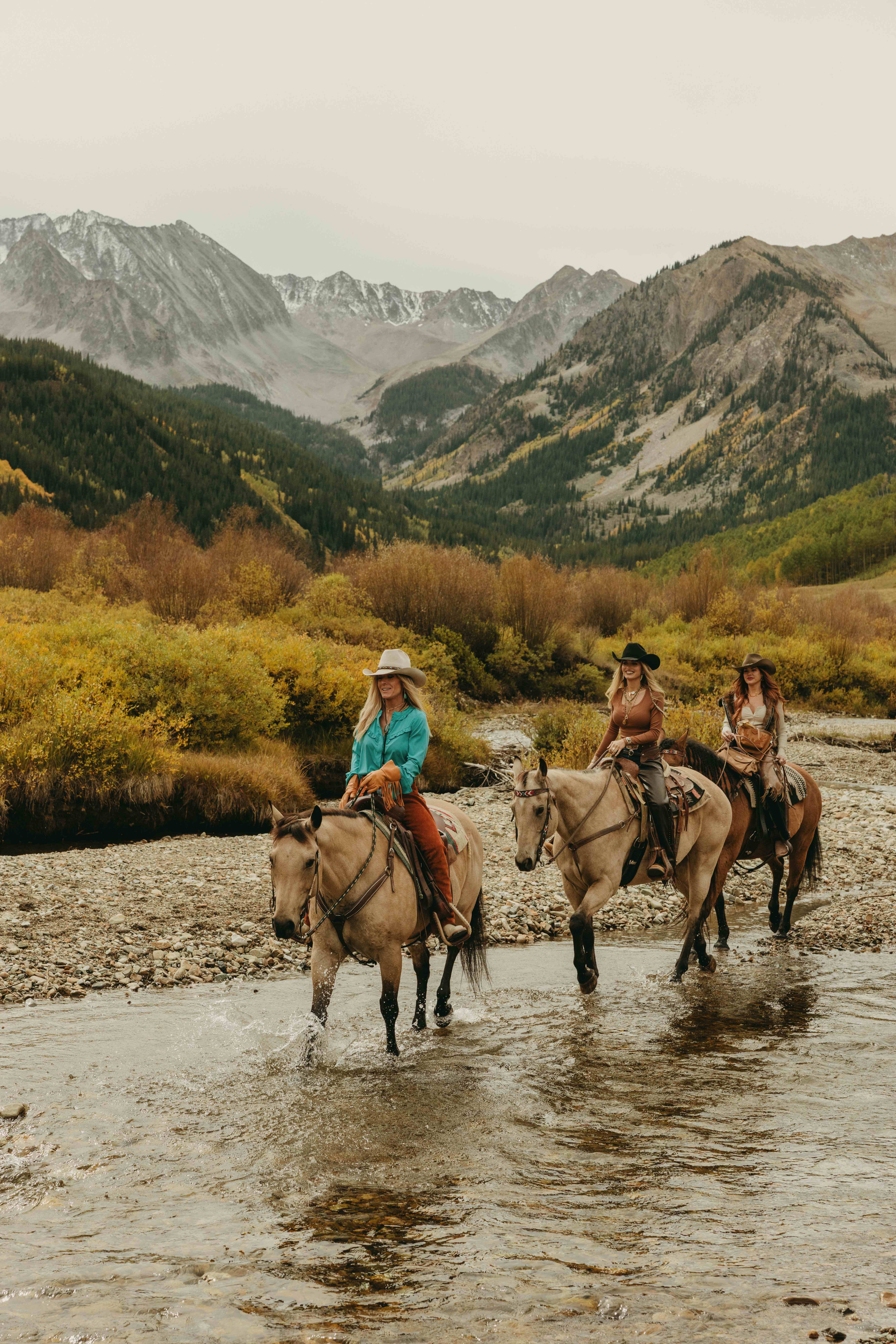 Three women riding horses through river with mountain backdrop