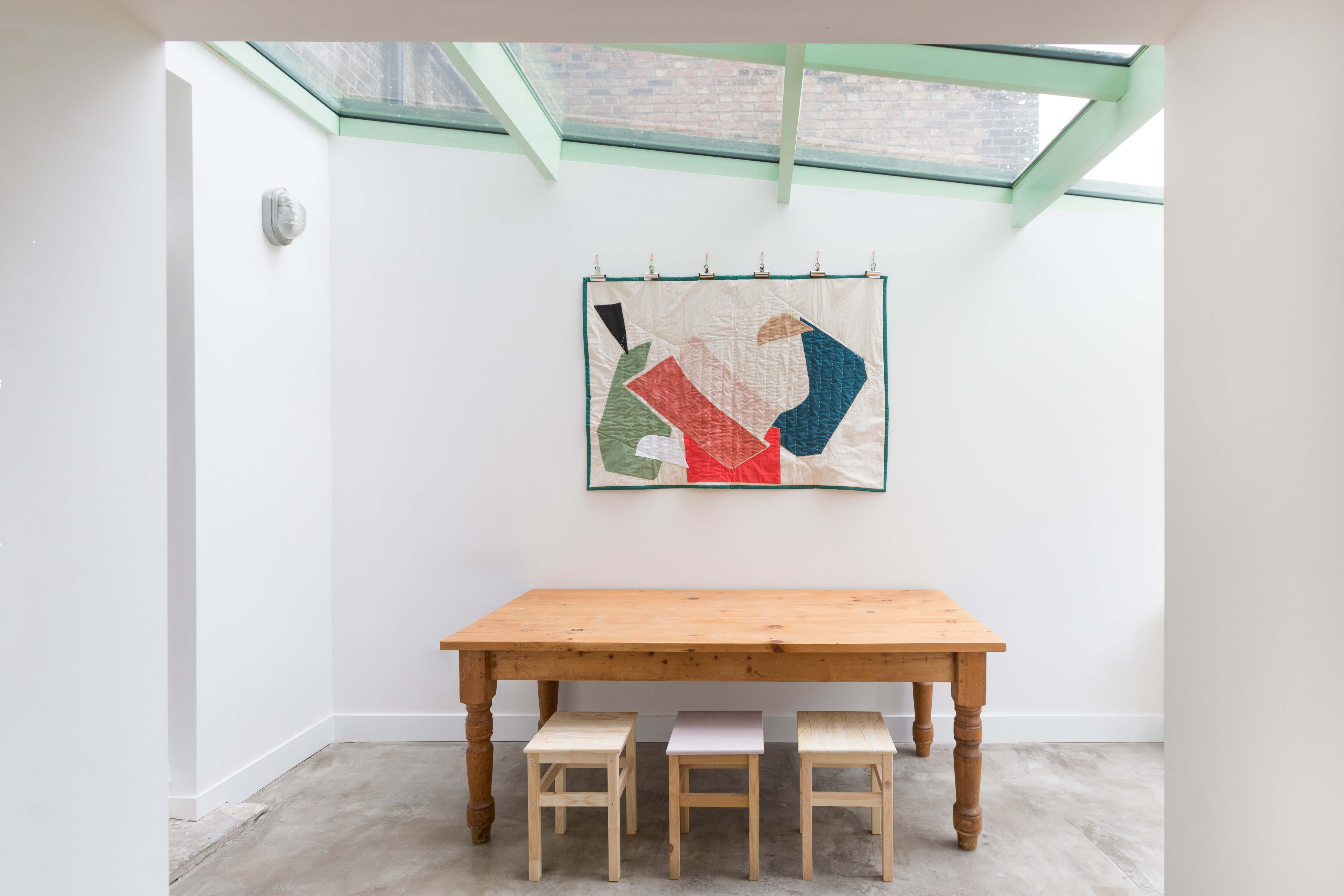 Minimalist dining area under glass roof with wooden farmhouse table, stools, and contemporary textile art